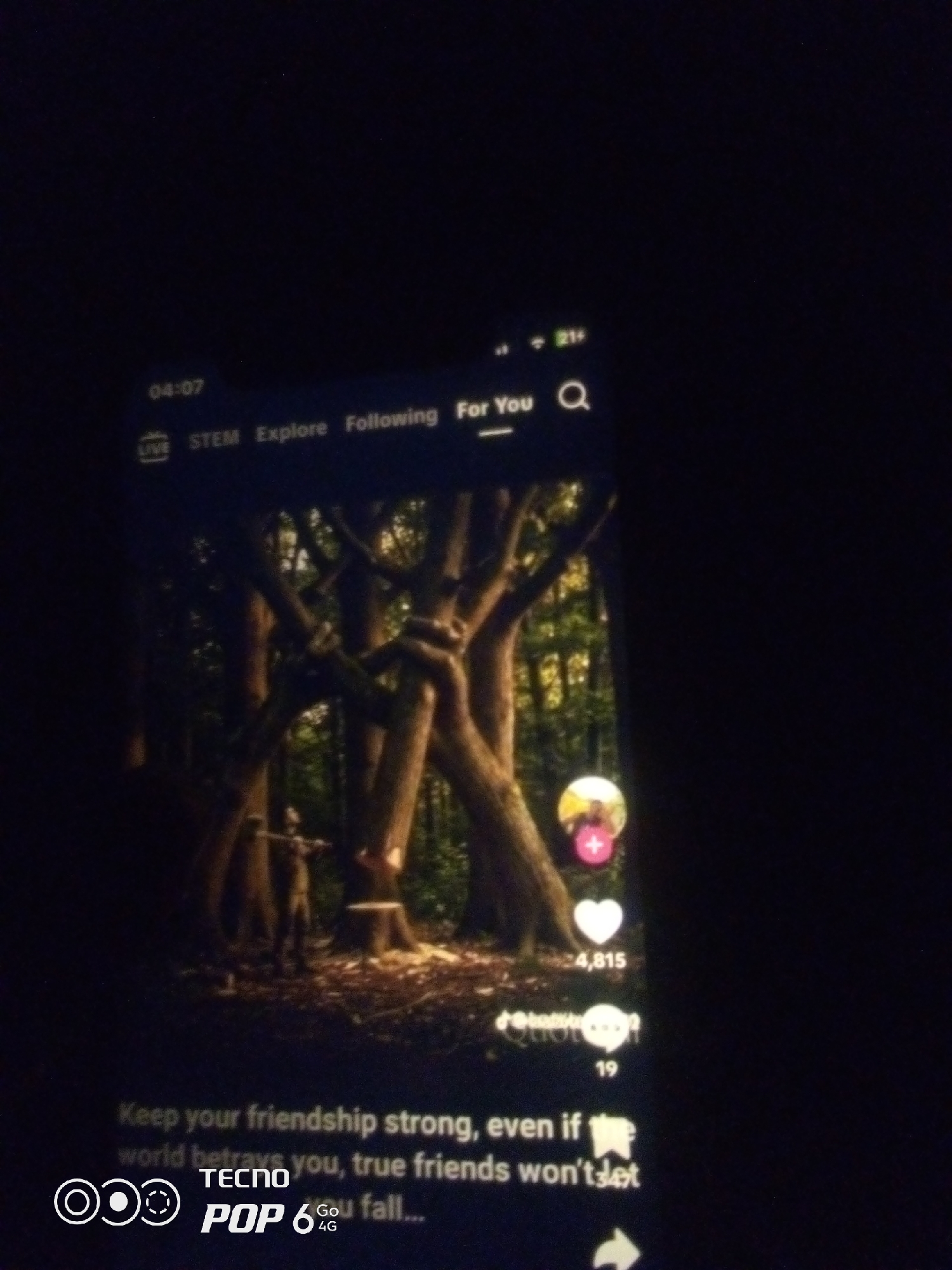 The image is a low-light photograph of a smartphone screen displaying a TikTok video. The video features a person standing in a forest with large trees. On the screen, the time "04:07" is visible at the top left, followed by navigation options: "STEM", "Explore", "Following", and "For You". A search icon is also present. To the right of the video, there are interaction icons, including a profile picture with a pink plus sign, a heart icon with the number "4,815" next to it, and a speech bubble icon with the number "19". At the bottom of the screen, text reads: "Keep your friendship strong, even if world betrays you, true friends won't let you fall...". The bottom left corner of the screen displays the TECNO POP 6 4G logo. The overall scene depicted on the phone is a forest environment, but the image itself was captured in a dark room, with the phone screen being the primary light source. There is no information in the image to suggest a specific location within Howayi, Nigeria.
