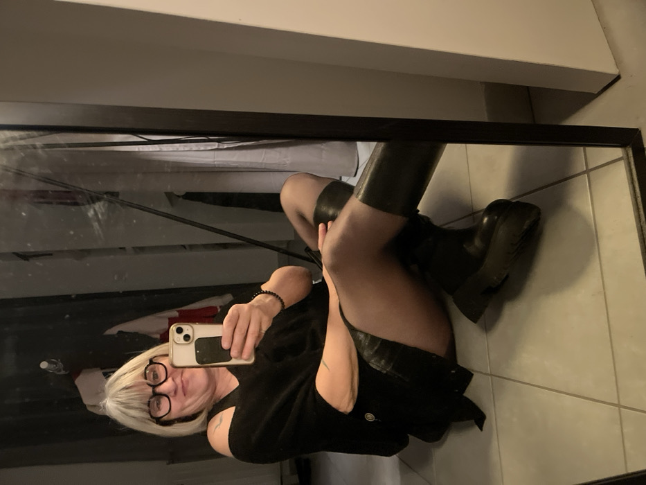 A mirror selfie captures a woman with blonde hair and glasses wearing a black dress, dark tights, and knee-high black boots. She holds a phone with a white case. The reflection reveals a room with a tiled floor and what appears to be a bed behind her. The mirror has a dark frame.