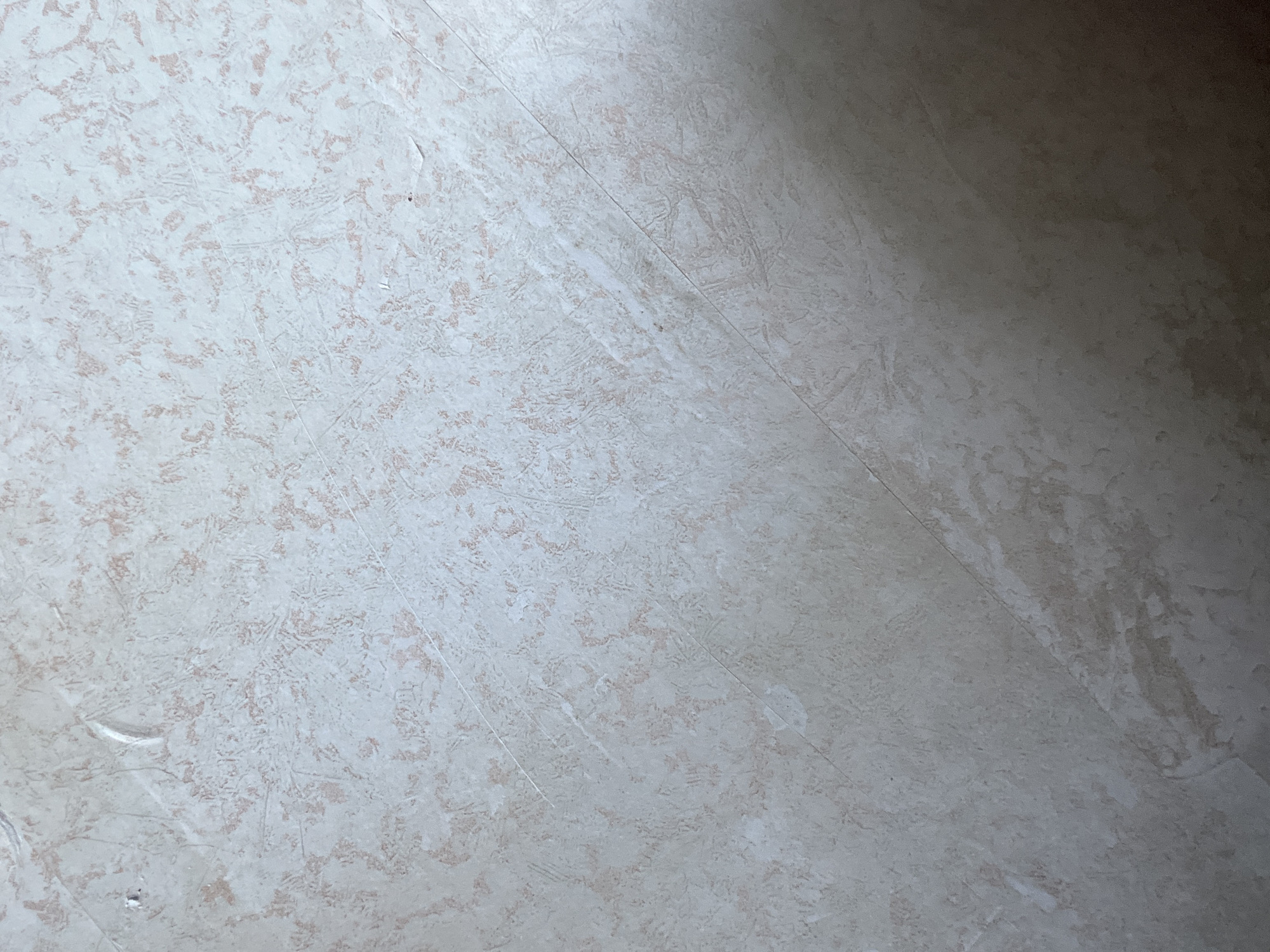 The image displays a close-up view of a textured, light-colored surface, possibly wallpaper or a painted wall, with subtle pinkish-beige speckles and faint diagonal lines that appear to be creases or seams. The lighting creates a shadow on the lower right portion of the image, indicating the direction of light is from the upper left. There are no discernible people, subjects, or actions. The location in the city of Красные Баки, Russia, is not visually represented in the image itself.