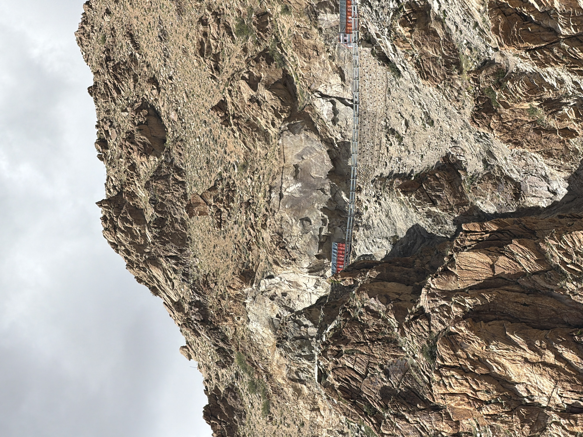 The image depicts a rugged, mountainous landscape with a road carved into the side of a steep incline. Two red and white trucks are visible on the road, one in the middle ground and another further in the distance on the right. The terrain is rocky and arid, with sparse vegetation. The sky is overcast, suggesting a cloudy day. The scene is located in the city of Leh, India, a region known for its dramatic mountain scenery.