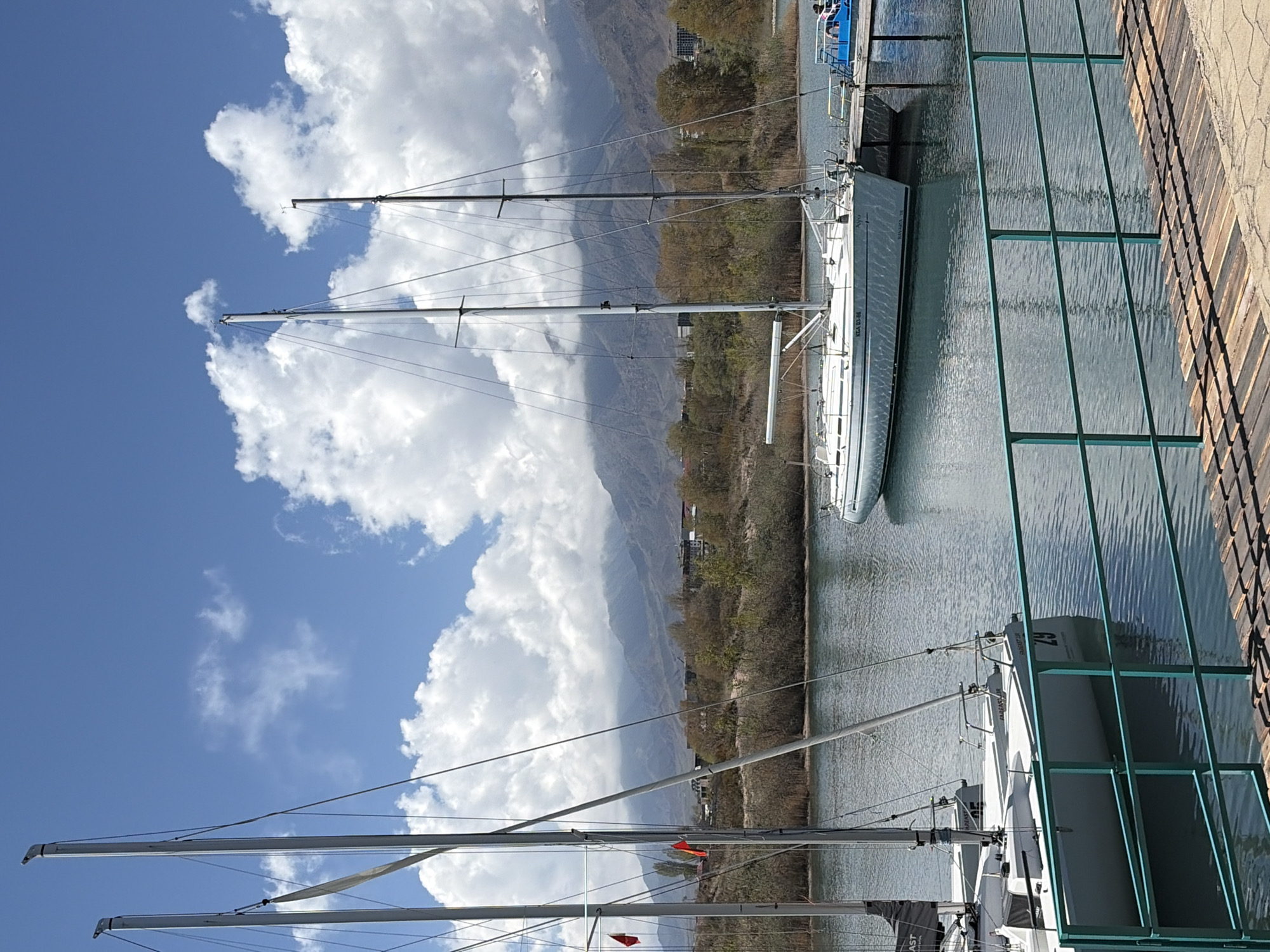 The image displays a serene marina scene, likely on a lake, under a bright blue sky with large, white cumulus clouds. In the foreground, a green metal railing or fence runs along a tiled or wooden walkway on the right. Several sailboats are docked in calm, rippling water.
The central sailboat is white with a tall mast and visible rigging. On its hull, the text "NEA 03-86" and "BAVARIA 30 cruiser" is clearly visible. Part of another sailboat is seen in the bottom left. Beyond the docked boats, the distant shoreline is lined with sparse trees and some buildings at the base of large, hazy mountains. The mountains dominate the background, with some of their peaks shrouded by the bright white clouds.
The scene conveys a peaceful, clear daytime atmosphere. No people are visible. The location, Çolpon-Ata, Kyrgyzstan, suggests this is a view of Lake Issyk-Kul, surrounded by mountains.