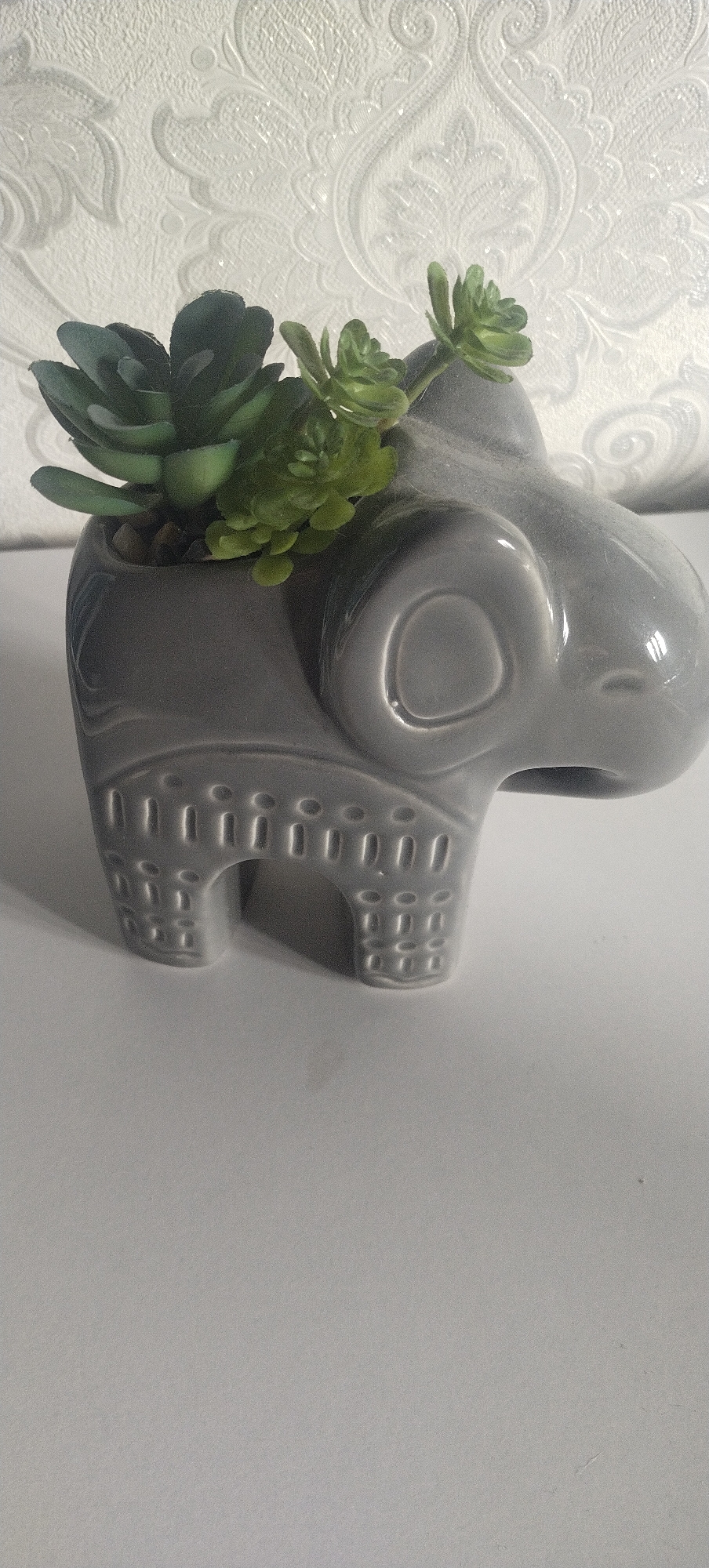 A close-up shot of a gray elephant-shaped ceramic planter sits on a white surface, against a white textured wallpaper. The planter is filled with artificial succulents, two dark green and two lighter green. The planter has raised circular and rectangular patterns on its sides and front legs, resembling decorative motifs. The background wallpaper has a raised damask pattern. The scene is brightly lit, with soft shadows cast on the white surface below. There is no indication of location in the city of Unknown, Unknown.