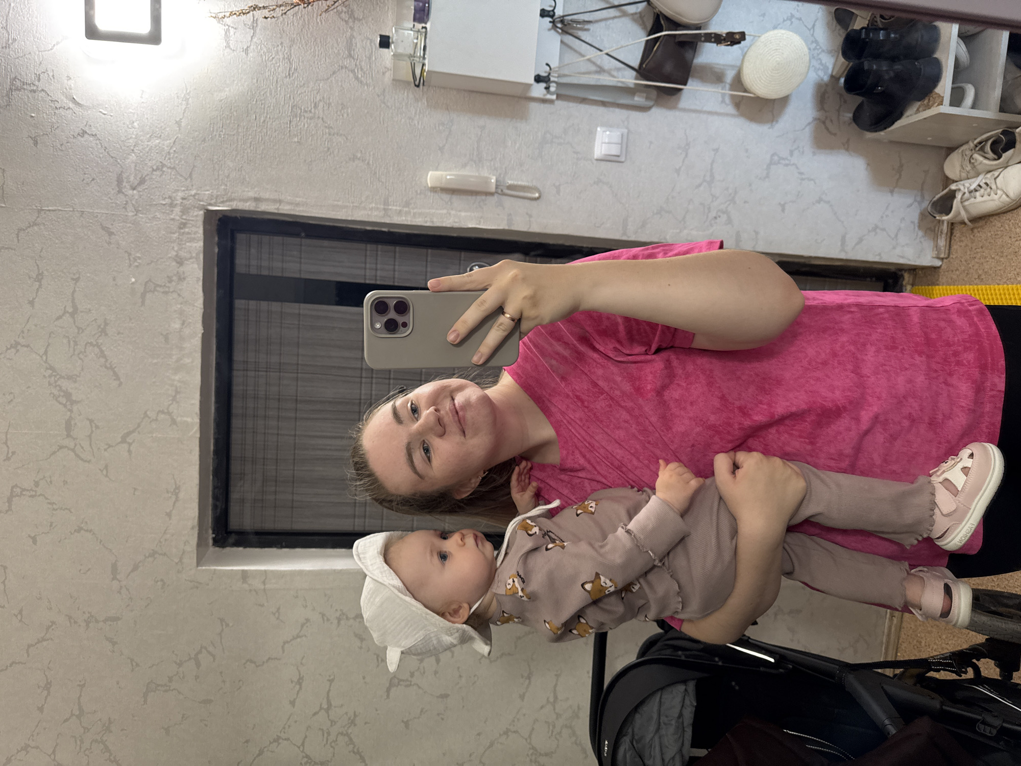 A woman is holding a baby while taking a selfie. The woman is wearing a pink shirt. The baby is wearing a beige outfit with fox patterns and a white bonnet. The baby also has pink sandals on. A black stroller is partially visible at the bottom. The setting appears to be inside a home, based on the wall texture and the presence of home objects like a clothes drying rack and shoes. It is not possible to confirm the location in Chaikovsky, Russia from the image.
