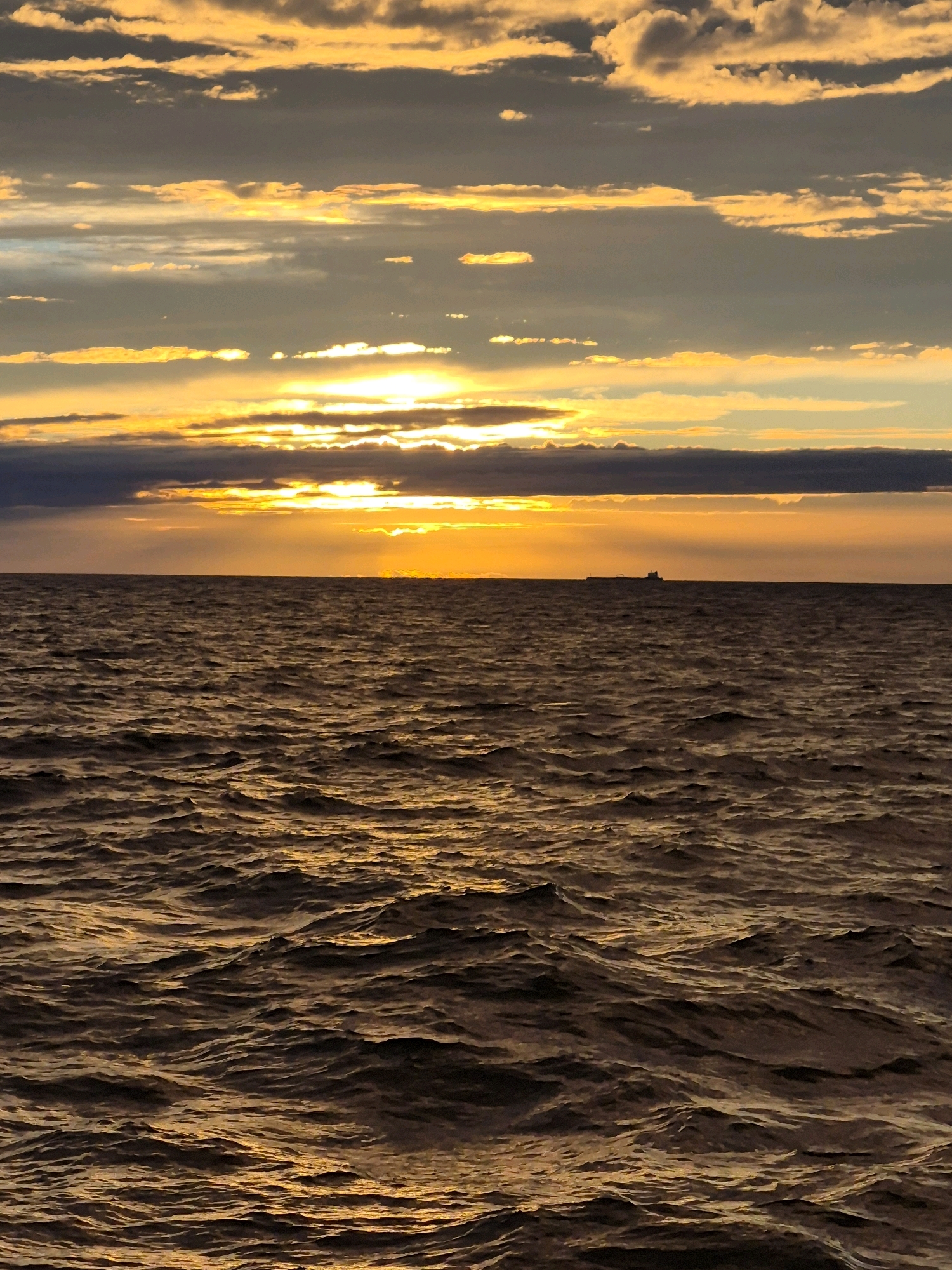 An outdoor, full-shot, eye-level, vertical, medium lighting photograph captures a sunset over the ocean. The sky is filled with clouds, illuminated by the setting sun in hues of orange and yellow. The sun is partially obscured by a band of dark clouds near the horizon, with rays of light piercing through. The ocean stretches out in the foreground, with gentle waves reflecting the warm colors of the sky. A faint silhouette of a cargo ship can be seen on the horizon. The overall scene evokes a sense of calm and tranquility. The image is a photograph of an ocean sunset, with no discernible urban features or identifiable city locations.