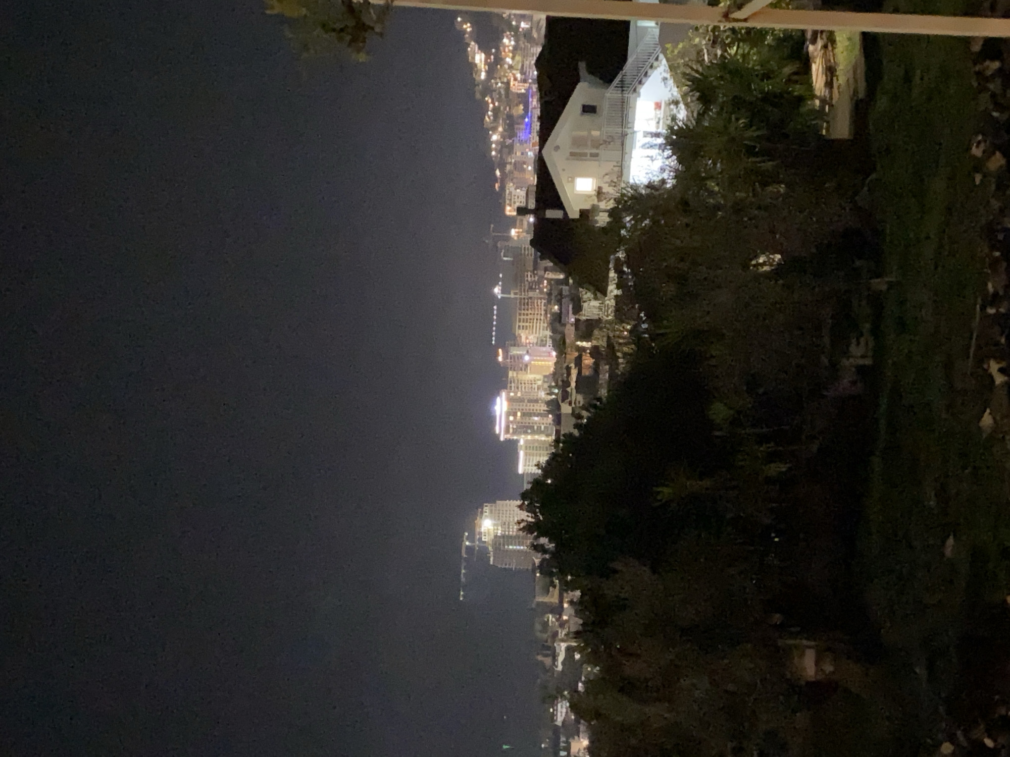 The image depicts a nighttime view of Budva, Montenegro, from an elevated vantage point. The illuminated cityscape stretches along the coast, with tall buildings and various light sources creating a vibrant urban scene. In the foreground, trees and foliage are present, partially obscuring the view. A white building is situated near the viewing point. The sky above the city is dark.