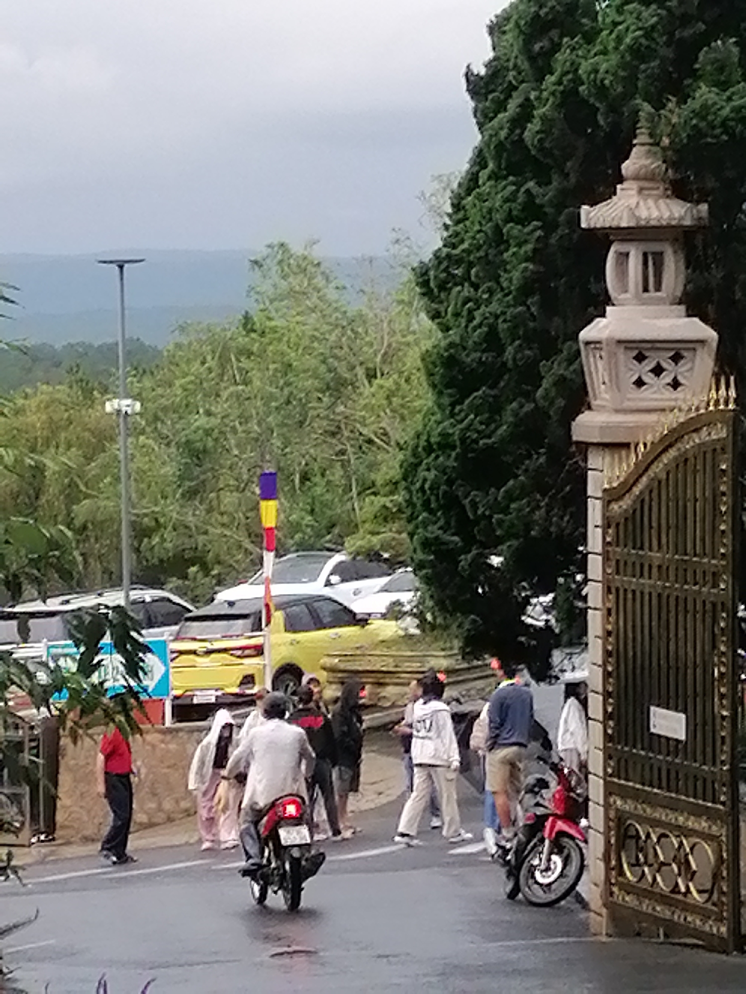 The image shows a street scene in Da Lat, Vietnam, based on the Asian-style architecture of the gate and the surrounding lush, hilly landscape. People are gathered near an entrance with an elaborate golden gate and a stone lantern. A man on a motorcycle rides away from the group. Other individuals are standing or walking nearby. Behind them are parked cars, including a distinctive yellow vehicle. A tall, modern streetlight is visible. The overall impression is of a cloudy, possibly rainy day.