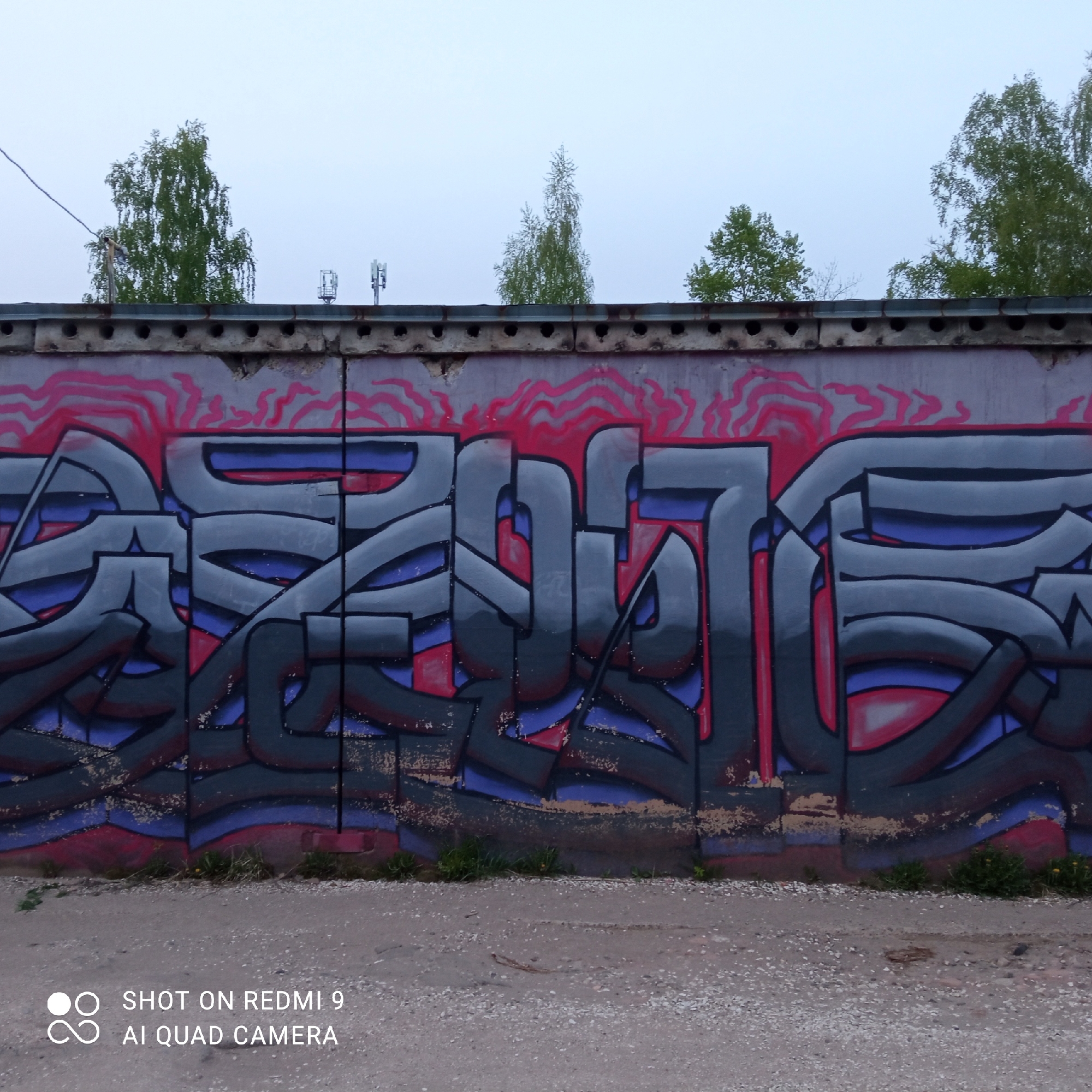 The image depicts a concrete wall with graffiti in Chaikovsky, Russia. The graffiti is a stylized, abstract design using black, gray, purple, and red paint. Above the graffiti is a row of concrete blocks with circular holes. Above the blocks, sparse trees and two antennae or towers can be seen against a light, overcast sky. At the base of the wall, there is a gravel and dirt ground with sparse weeds. The bottom left corner of the image shows a watermark indicating "SHOT ON REDMI 9 AI QUAD CAMERA".