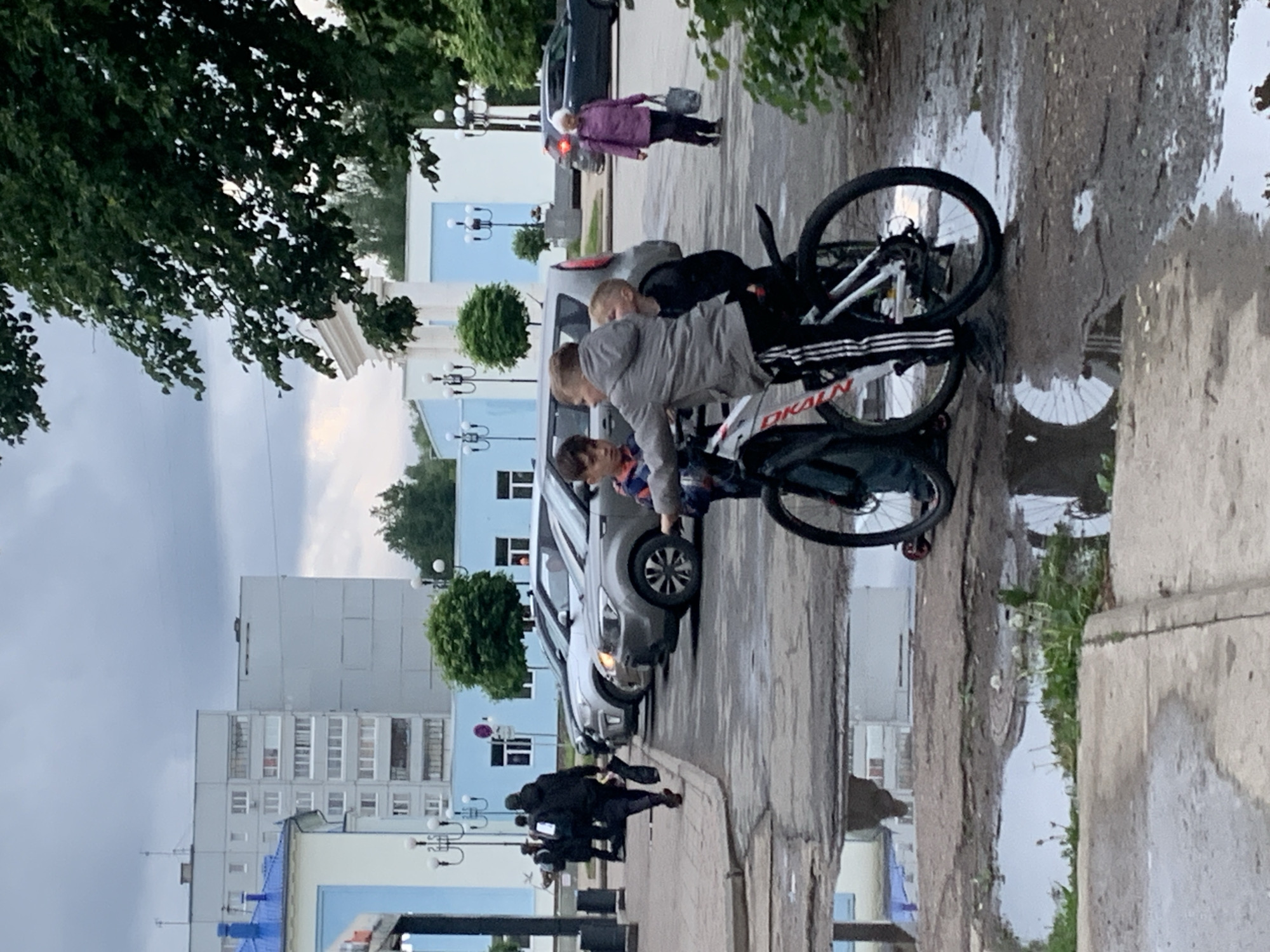 The image shows a street scene in what appears to be the city of Chaikovsky, Russia, evident from the text "Чайковский" visible on the bicycle frame. The day appears to be overcast and wet, with puddles reflecting the surroundings.
In the foreground, there is a bicycle parked on the sidewalk, a group of people clustered around it, possibly children and an adult. To the right, a puddle reflects the bicycle and surrounding elements, distorting the image.
Beyond the bicycle and group, there are parked cars, and further back, pedestrians walking on the sidewalk. The background features buildings, including multi-story apartment blocks and lower, possibly administrative or commercial buildings painted in light blue with classical architectural details. Street lamps are visible. Vegetation includes trees lining the street.