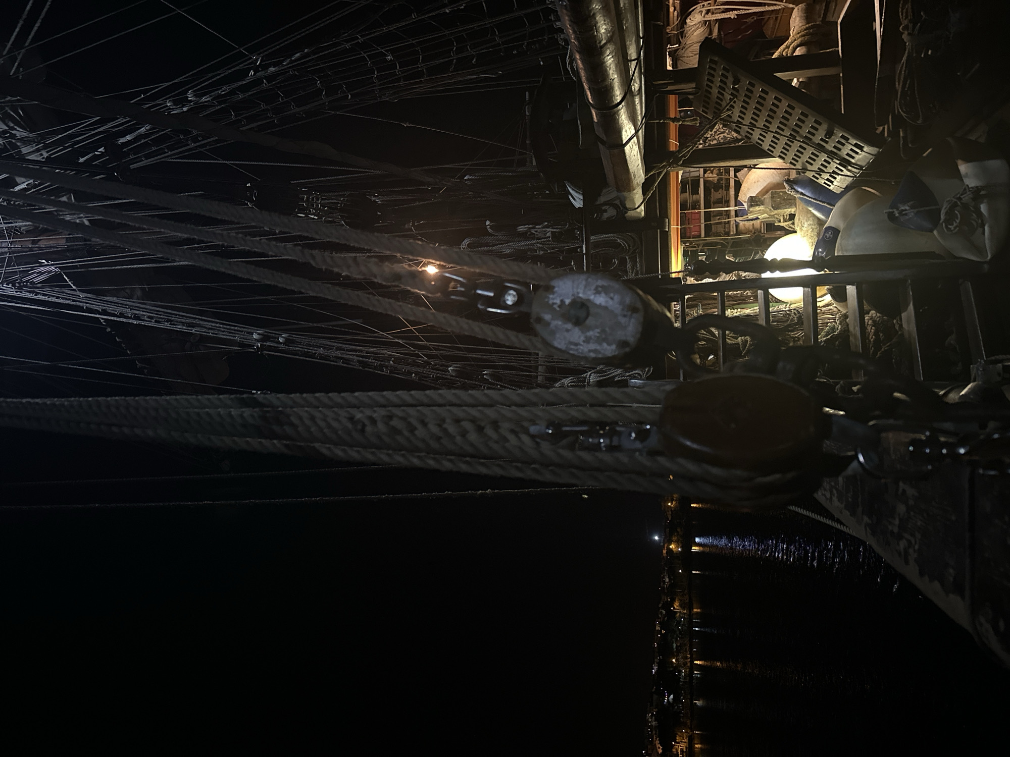 The image depicts the rigging of a tall ship at night, likely the "Çamlık" docked in Datça, Turkey. The photograph is taken from a low angle, looking upward into the complex network of ropes and spars. Two prominent wooden blocks are visible in the foreground, with thick ropes running through them. Above, a tangle of thinner ropes and lines fills the frame, partially illuminated by a light source further up the mast. To the right, the ship's structure is visible, with metal grates and other equipment in view. The water reflects some of the surrounding city lights.