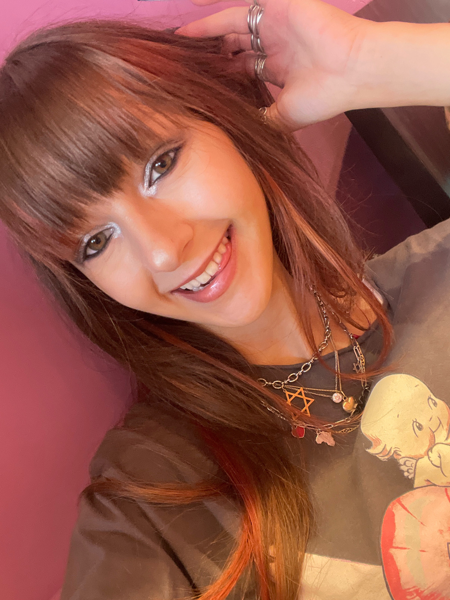 The image is a close-up selfie of a young woman with long, brown hair streaked with reddish-orange highlights. She is wearing a dark gray t-shirt with a cartoon image on it, and several layered necklaces, including one with a Star of David pendant. Her eyes are lined with silver eyeshadow, and she has her tongue sticking out slightly. Her left hand is raised, touching her hair, and she is wearing several silver rings on her fingers. The background is a solid pink wall, suggesting an indoor setting. There is no visual information in the image to determine its specific location within Raanana, Israel, or any other city.