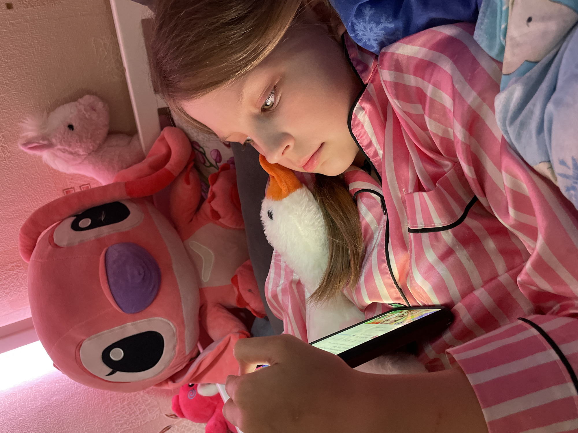 A young girl, appearing to be around 8-10 years old, is lying in bed, looking at a smartphone. She is wearing pink and white striped pajamas with black trim. She has a fair complexion and light brown hair. Her head is resting against a white stuffed animal goose with an orange beak.
Surrounding her are several other stuffed animals, including a large pink plush toy with a purple eye, and a smaller pink stuffed animal in the background. The bedding includes a blue blanket with a snowflake pattern. The room has light-colored walls. There is no information indicating the scene is located in Izhevsk, Russia.