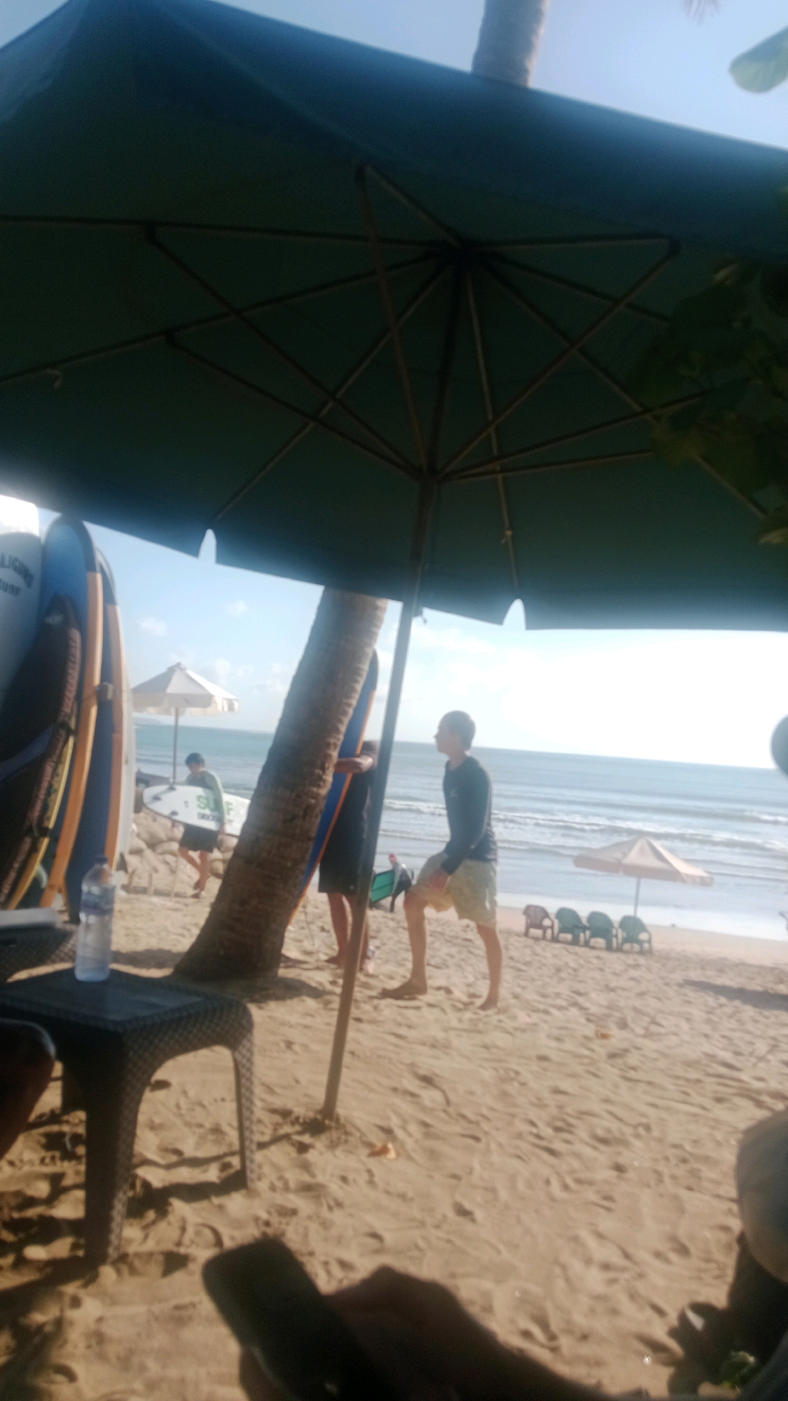 The image depicts a beach scene in Kuta, Indonesia. A large, dark green umbrella covers the foreground, casting a shadow over the sandy beach. To the left, several surfboards are propped up, and a plastic table with a water bottle on it is visible. A person is partially in view on the left, sitting and holding a phone. In the middle ground, a person stands near a palm tree, and another person is visible in the background near the ocean. Several beach chairs and a small umbrella are set up on the sand. The ocean with gentle waves stretches to the horizon under a clear sky. The overall atmosphere is relaxed and sunny, characteristic of a beach resort town.