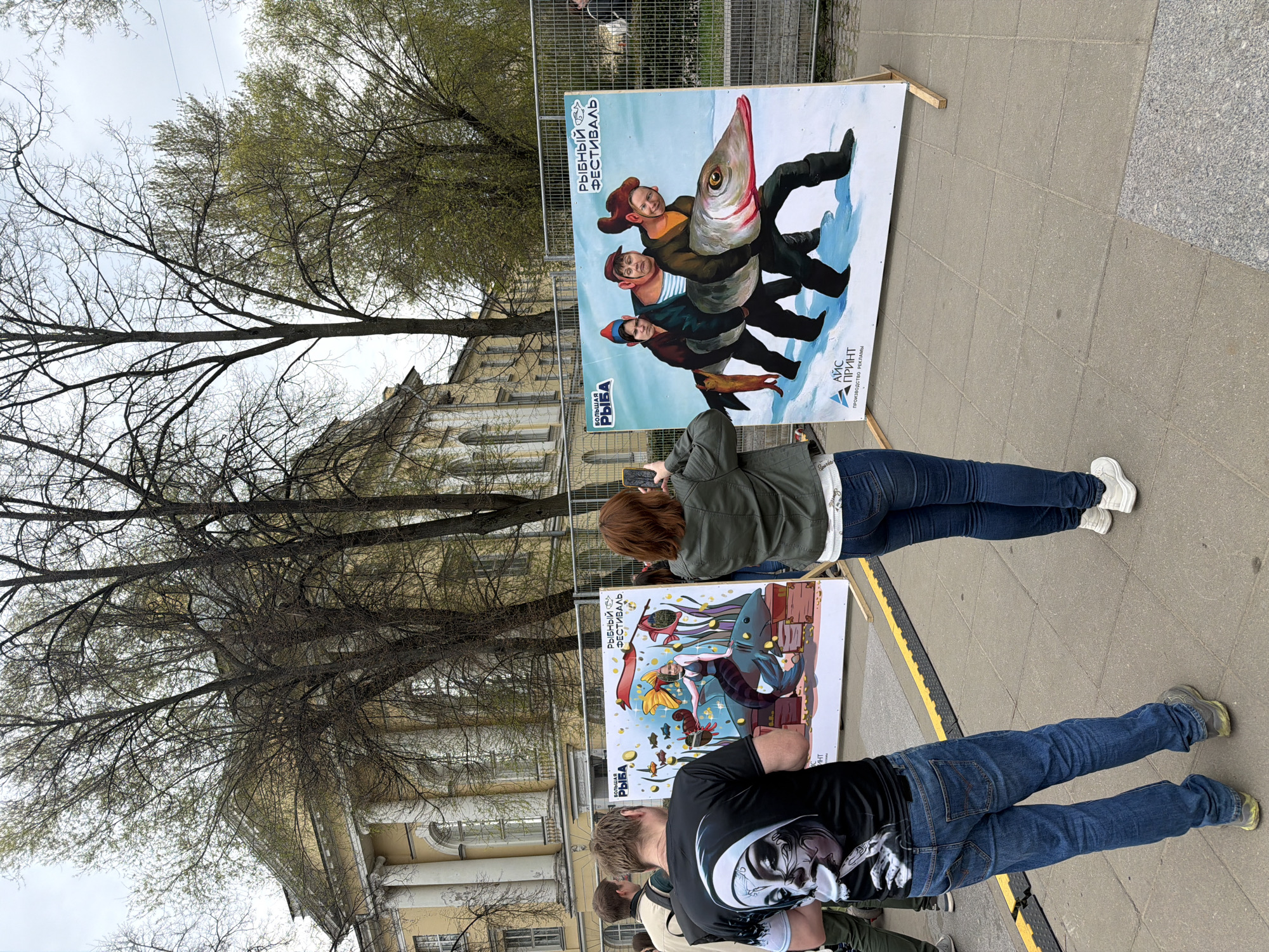 The image shows an outdoor scene in Saint Petersburg, Russia, with two large display boards propped up on a sidewalk. The boards appear to be promoting a fish festival, displaying colorful illustrations.
A woman stands in front of the upper board, holding a mobile phone and taking a picture of the artwork. She is wearing a green jacket, jeans, and white sneakers. Next to the lower board, a man is standing, clad in a black T-shirt with a graphic print, and blue jeans. Several other people are partially visible to the left of the man.
Behind the boards, a building with light-colored facade and visible windows can be seen, along with bare trees. The sky is overcast. A metal fence runs along the edge of the sidewalk. The environment suggests an urban setting with cultural or promotional displays.