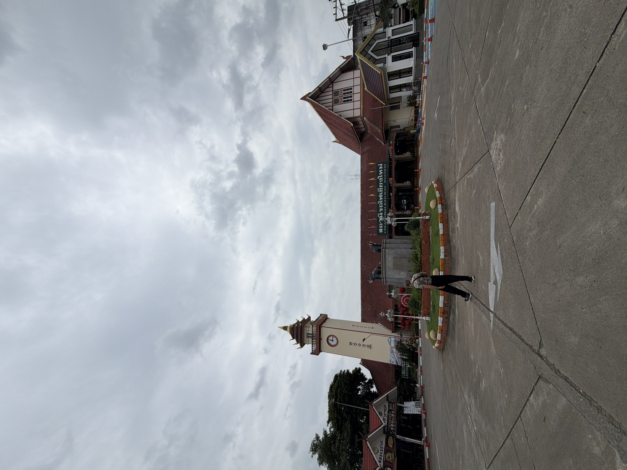 A person walks across a paved area in front of the Chiang Mai Railway Station in Chiang Mai, Thailand. The station building has a distinctive clock tower with a traditional Thai roof and intricate architectural details. The main station building is visible to the right of the clock tower, also featuring a red tile roof. In the foreground, a circular landscaped area with decorative stones is present. The sky is overcast with clouds.