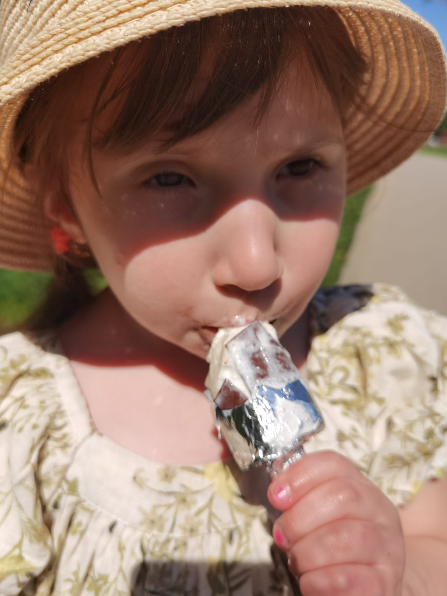 A young child, with bangs and a straw sun hat, is holding and eating an ice cream bar wrapped in foil. The child is wearing a light-colored dress with a floral pattern. The location is presumed to be Perm, Russia, but that cannot be determined from the image alone.