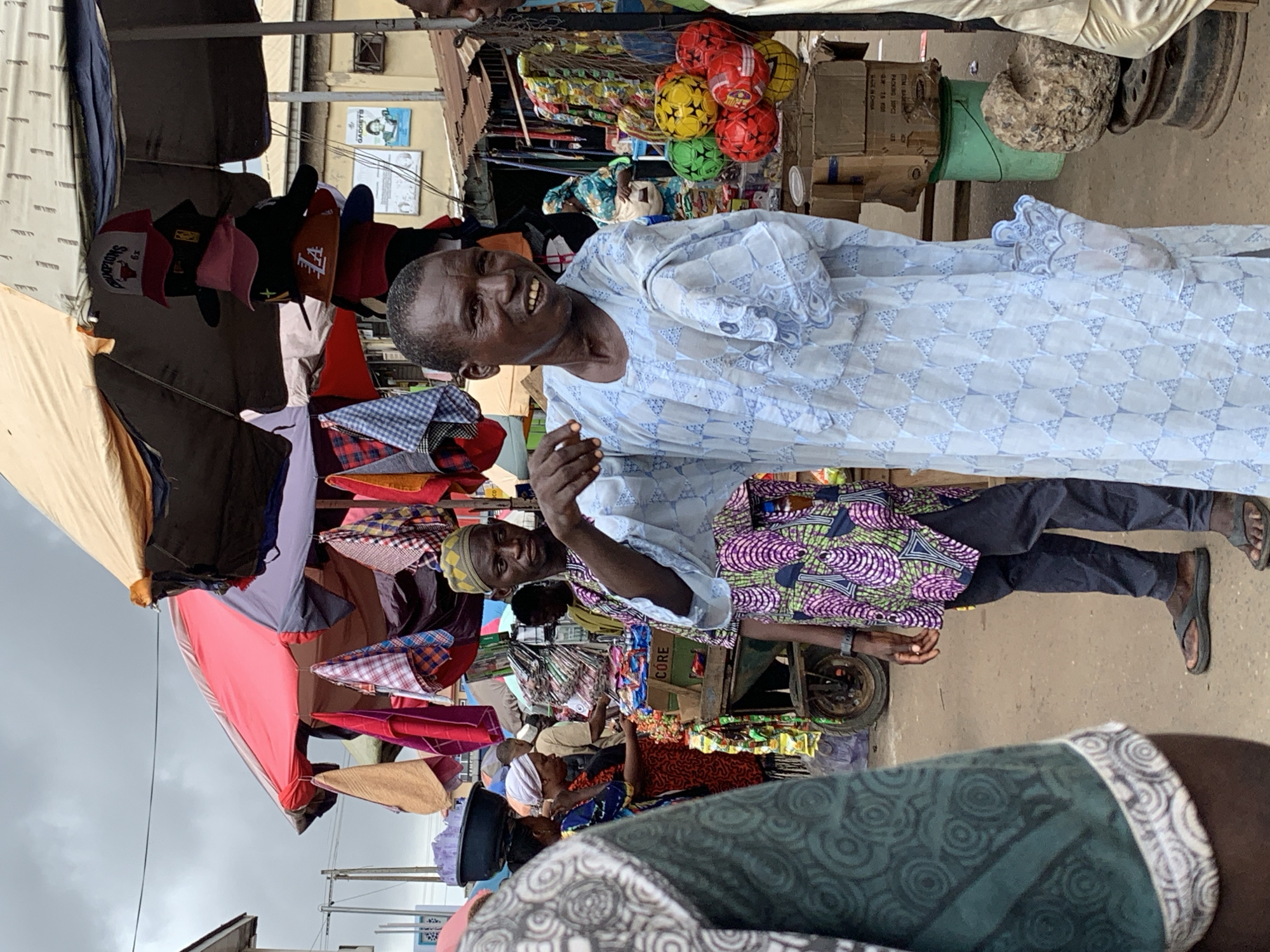A street scene in Ikorodu, Nigeria, shows two men, one wearing a light blue patterned robe, smiling and gesturing toward the camera. The other man wears a colorful shirt and dark pants. They stand amidst a bustling marketplace with vendors and shoppers. Merchandise, including hats, fabrics, and balls, are displayed under makeshift canopies. A cart filled with goods is visible in the background. Overhead power lines and the sky suggest an outdoor setting.