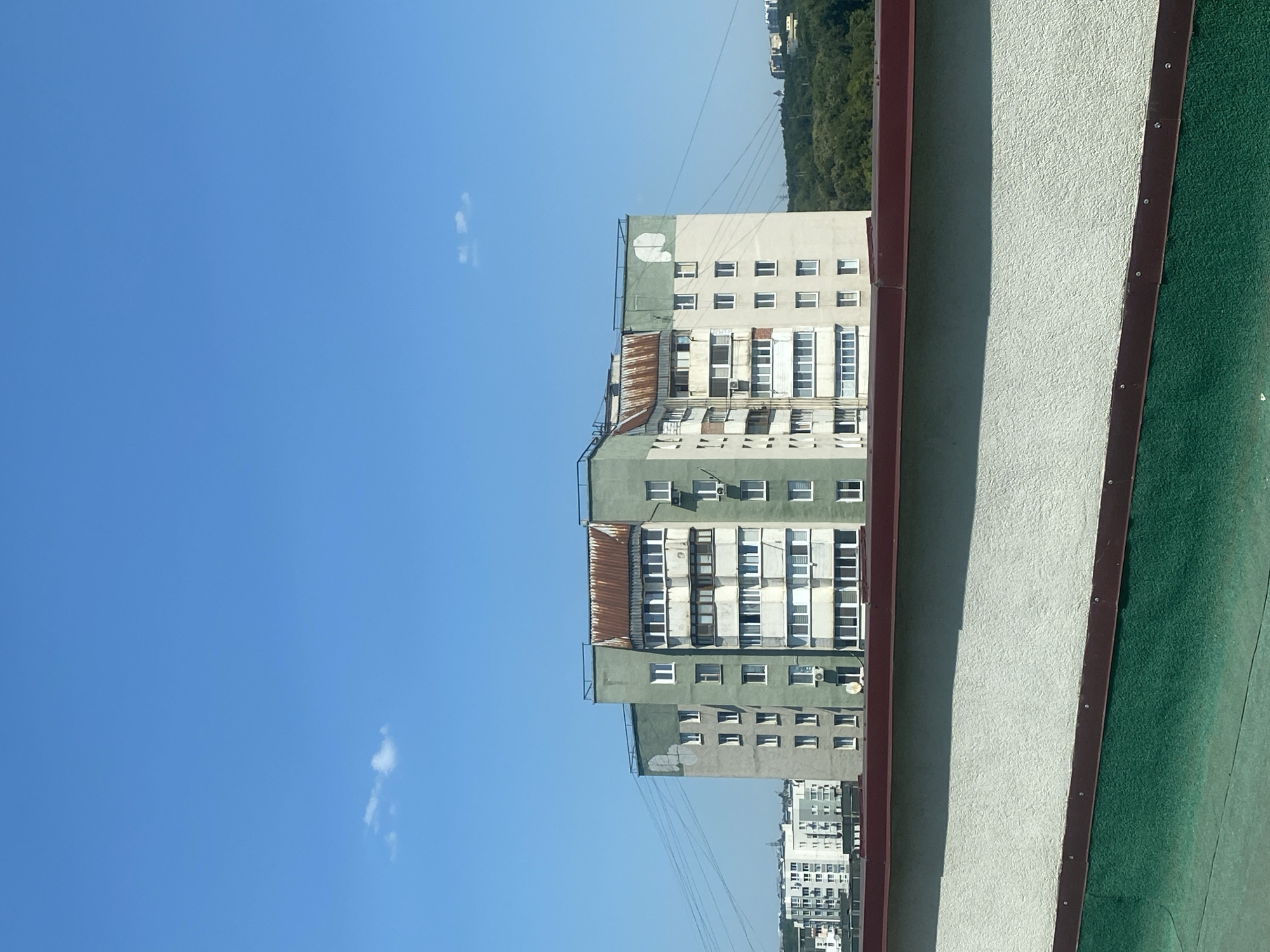 The image shows a multi-story apartment building with a green and white facade, some sections of which appear weathered and stained. The building has numerous windows, some with balconies. The roof of the building has sections covered with what appears to be rusted corrugated metal. In the foreground, there is a parapet with a textured white surface and a dark red trim, below which is a dark green surface. Several utility wires are visible, extending from the building towards the left. In the background, other buildings and a dense area of green trees are visible under a clear blue sky with a small, wispy cloud. The perspective suggests the photo was taken from an elevated position, possibly another building or rooftop. The style of the apartment building is typical of Soviet-era architecture, commonly found in Eastern European cities. Without specific identifying landmarks, it is impossible to pinpoint the exact location within Chișinău, Moldova. The scene depicts a residential urban environment on a clear, sunny day. There are no people visible in the image.