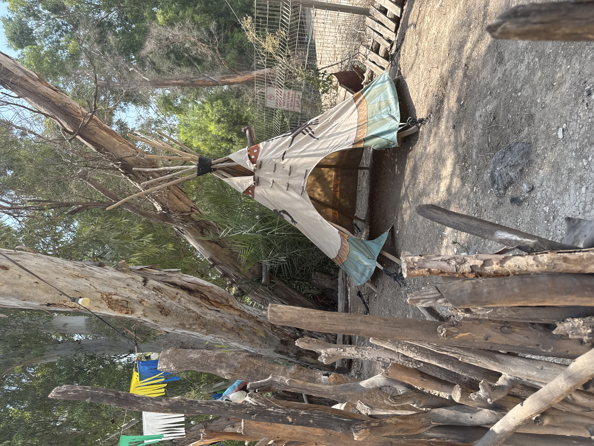 The image depicts an outdoor scene at a campsite or recreational area within the Emek HaYarden Regional Council, Israel. In the foreground, a rustic fence constructed from irregularly shaped wooden poles is visible, creating a sense of enclosure. Behind this fence, a teepee-style tent is set up, its fabric decorated with geometric patterns in earthy tones and green. The tent is supported by wooden poles and appears to be an open structure. Large trees with rough bark dominate the background, with foliage suggesting a dry, possibly arid environment. A string of lights hangs from one of the trees, and colorful fabric banners are also strung between trees, indicating a festive or decorated atmosphere. A metal fence with a "no entry" sign is visible on the right side of the frame. The ground is covered in dirt and small stones.