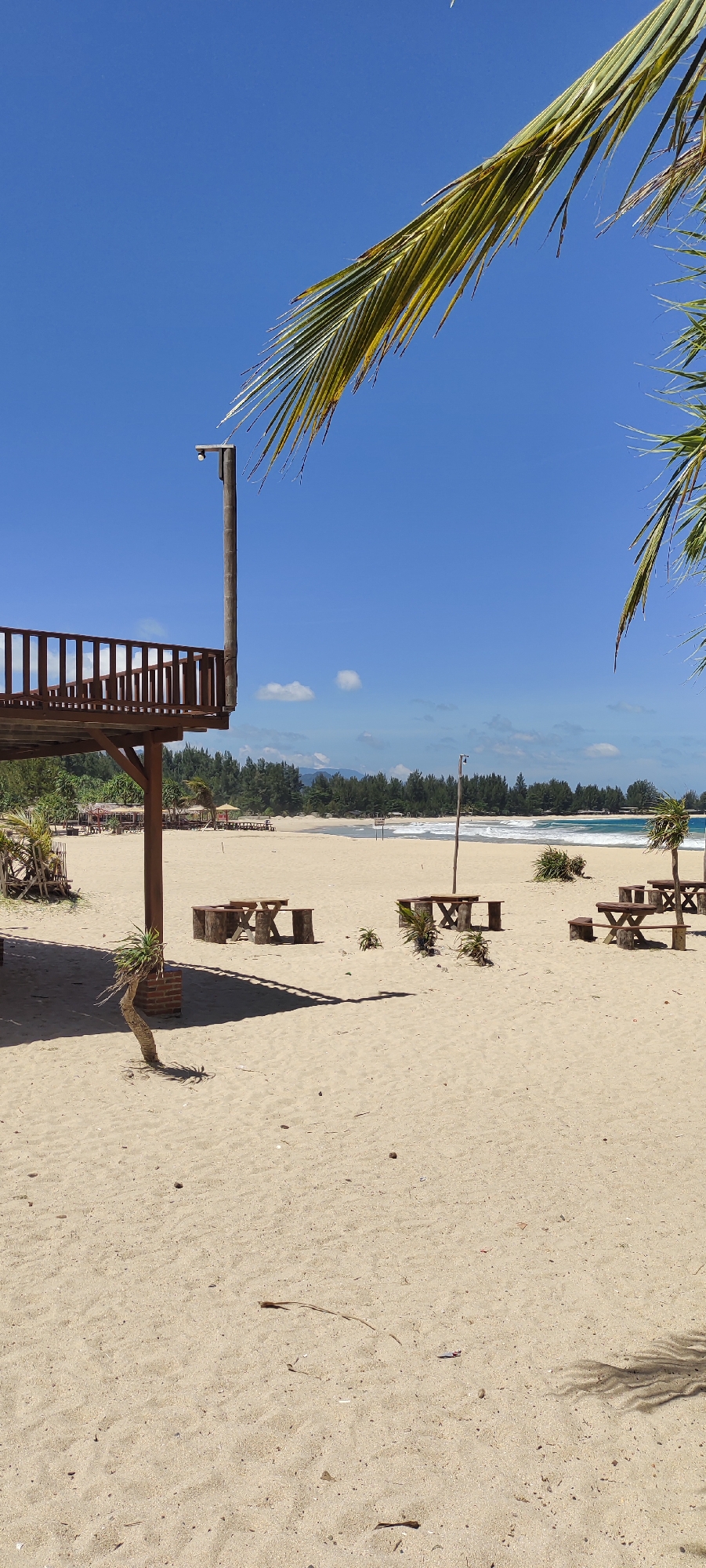 This is a photograph of a beach scene in Meunasahbale, Indonesia, on a clear, sunny day. The foreground is dominated by light-colored sand with some scattered debris. A wooden structure with a railing, likely a viewing deck or part of a beachside establishment, is visible on the left, supported by wooden posts. A small, thin tree with sparse foliage is growing near one of the posts.
Further into the scene, several picnic tables made of wood are set up on the sand. Some of these tables have benches attached. Behind the picnic area, a line of lush green trees forms a dense border along the coastline. The ocean is visible beyond the trees, with small waves breaking on the shore. The water is a vibrant blue, and the sky is a clear, bright blue with a few wispy white clouds. A single light pole with a camera or sensor attached stands near the wooden deck. Palm fronds from unseen trees hang over the top edges of the frame, indicating the tropical location.
