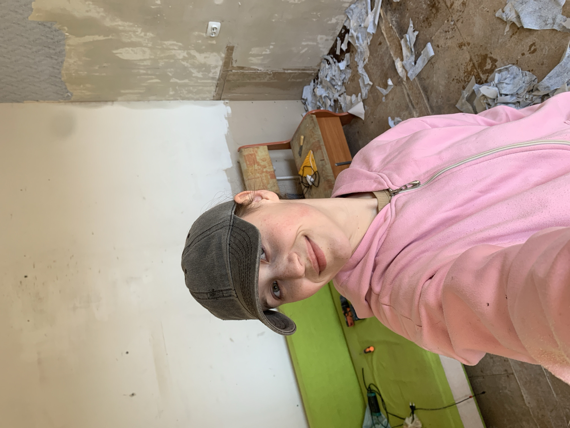 The image shows a person wearing a pink hooded sweatshirt and a gray cap, likely in a room under renovation. The room has partially stripped wallpaper, revealing the underlying wall surface. The floor is covered with debris, likely remnants of the removed wallpaper. A wooden table or shelf unit is visible in the background, along with a green object that appears to be a mattress. There is no indication of a city or specific location in the image. It's unlikely the location is in Фоки, Russia, as there is no information in the image indicating so.