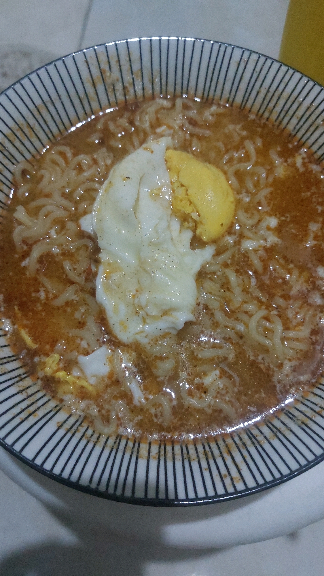 A close-up, overhead shot of a bowl of instant noodles with a cooked egg. The noodles are submerged in a reddish-brown broth. A partially cooked egg, with visible white and yolk, sits atop the noodles. The bowl has a black and white striped pattern on its rim. The scene is devoid of people and appears to be indoors, with a light-colored surface visible in the background. This image does not provide any information about its location in Jatirogo, Indonesia.