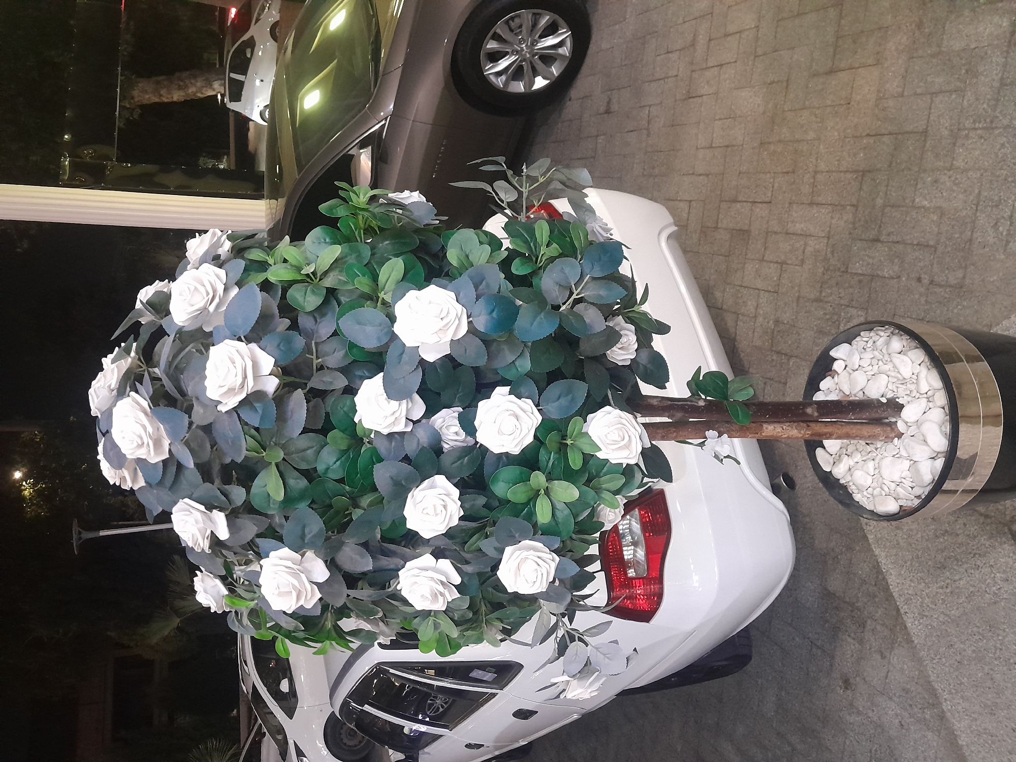 An outdoor, medium shot captures a decorative artificial floral arrangement of white roses and dark green leaves, placed in a black pot filled with white pebbles. The floral arrangement is positioned behind the rear of a white sedan, partially obscuring the car's taillight and trunk. The white sedan is parked on a paved surface, possibly a driveway or street, adjacent to a darker-colored sedan. In the background, there are trees and building structures, with evidence of artificial lighting due to the low-light conditions, suggesting it is nighttime. The composition is focused on the floral decoration and the vehicles.