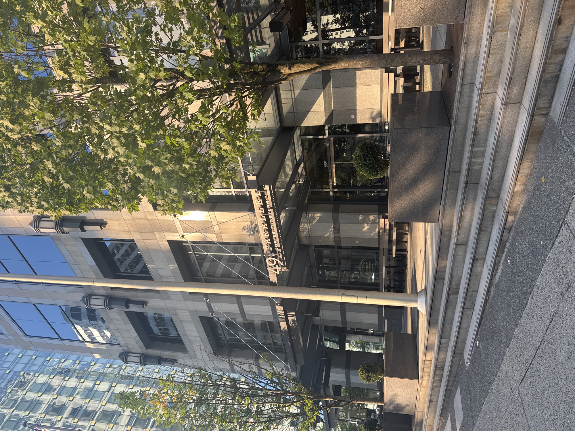 The image is an eye-level, outdoor, daytime shot of the building at 49 Parallel Coffee Roasters in Vancouver, Canada. The building is a modern structure with a glass and grey stone facade. Windows reflect other buildings in the area. An ornamental tree partially obscures the building. There are stairs leading up to the building entrance. Sidewalk visible on the right side.