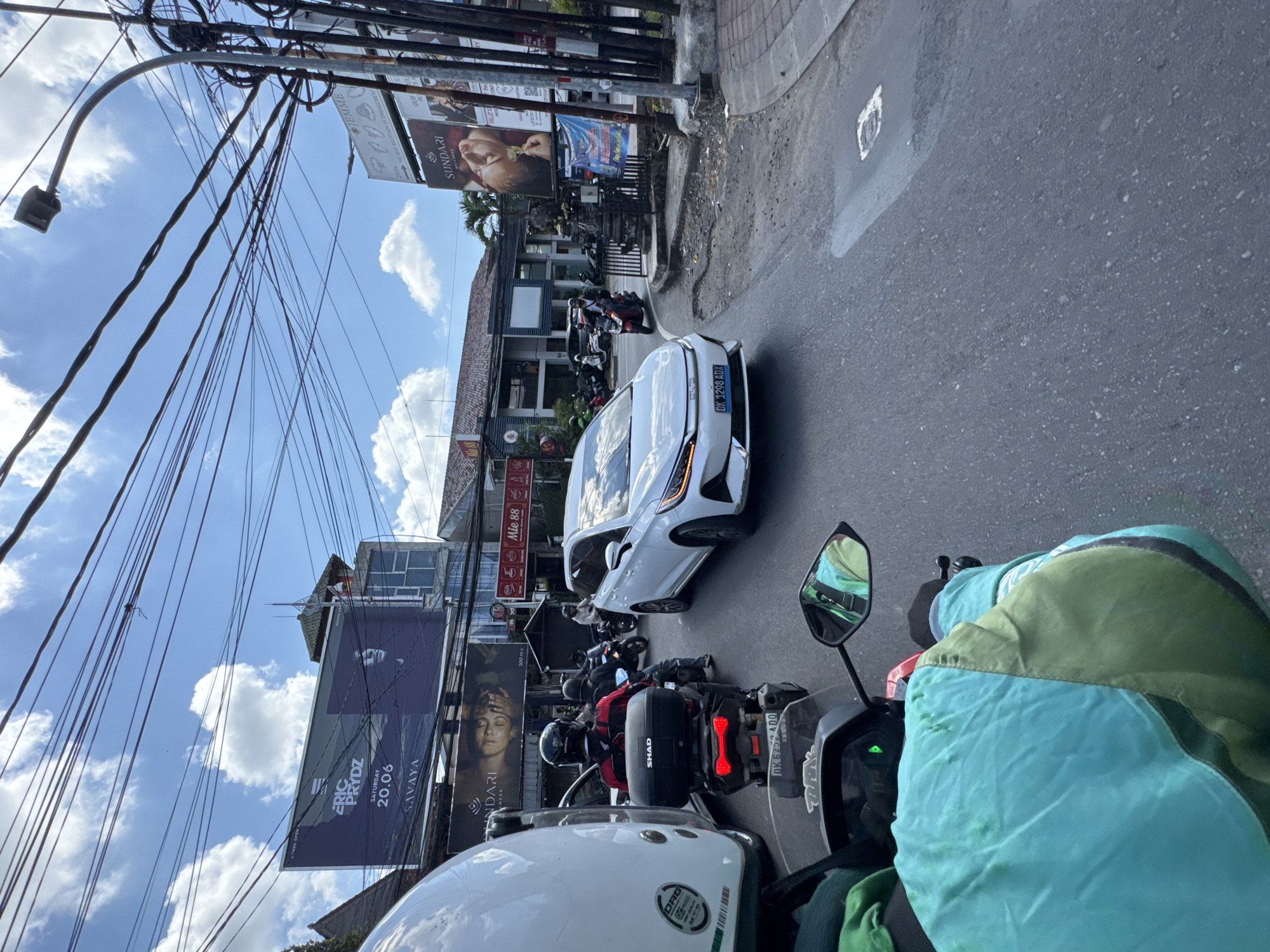 This image presents a bustling street scene in North Kuta, Indonesia, viewed from the perspective of someone on a motorcycle. The setting is a sunny day with a clear blue sky dotted with scattered white clouds.
In the immediate foreground, a white motorcycle helmet and a turquoise and green jacket belonging to the photographer are visible, along with parts of the motorcycle's dashboard and a side mirror. The main subjects are the various vehicles and people navigating the asphalt road. A white car, prominently featured, displays the Indonesian license plate "DK 9298 ADX," confirming its origin in Bali. Numerous motorcycles and scooters, many with riders wearing helmets, are present, either moving slowly or stopped in traffic.
The street is lined with buildings that appear to house commercial establishments. Overhead, a complex network of utility wires, typical of the region, crisscrosses the sky, supported by numerous poles which also hold street lights. Several large billboards and smaller signs are visible. A large billboard on the left advertises "ERIC PRYDZ SATURDAY 20.06 SAVAYA," indicating an event at a well-known club. Another partially visible billboard on the right features "SUNDARI" with an image of a woman's face, possibly for a spa or beauty product, and the text "sundari.ru." Other smaller signs include "MIE 88" and "WENKIE."
The activity captured is typical daily traffic and urban life, suggesting a busy period during the daytime. The overall impression is one of a vibrant, active street environment characteristic of North Kuta.