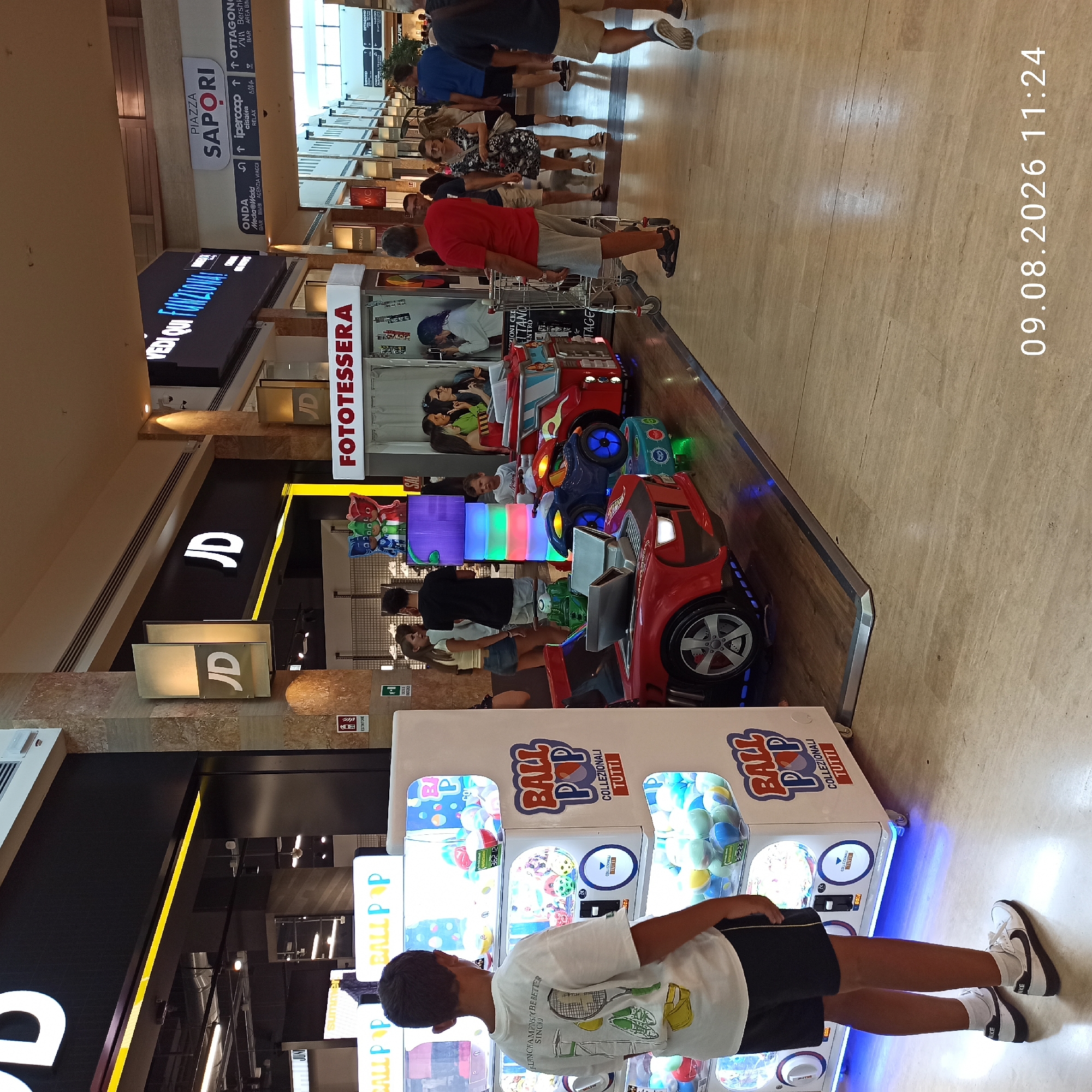The image depicts an indoor scene, likely a shopping mall in Molfetta, Italy, given the timestamp and context. In the foreground, a young boy with dark hair is seen from behind, wearing a white t-shirt with a tennis graphic and black shorts. He is standing in front of a "Ball Pop" claw machine, which is filled with colorful balls. Adjacent to the claw machine is a red children's amusement ride shaped like a race car. To the right of the car ride, a blue children's amusement ride resembling a motorcycle is visible. Further in the background, several adults are walking through the mall. To the left of the image, a store with a large "JD" logo is prominent, along with a smaller "JD" sign. Further down the mall, a large digital screen displays "VEDI QUI FUNZIONA!". Signs for "PIAZZA SAPORI" and various shops like "ipercoop" and "OTTAGONO" are also visible. The environment appears to be a well-lit, modern shopping center with tiled flooring.