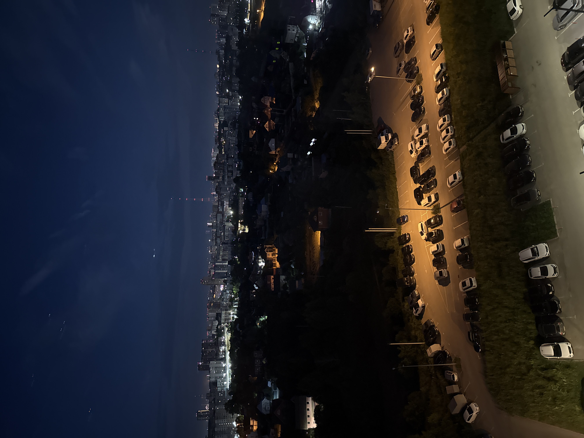 This is a night aerial photograph of a suburban or semi-urban area, likely on the outskirts of Perm, Russia, given the architectural style and the presence of apartment buildings in the distance. The scene is dominated by a sprawling cityscape illuminated by artificial lights, with a foreground of parked cars and houses. The sky is a deep indigo, with some faint clouds and a few distant lights that could be stars or distant aircraft.
In the mid-ground, a collection of houses, some with lit windows, are nestled among dark trees. Beyond the houses, a dense urban landscape stretches across the horizon, characterized by numerous apartment buildings of varying heights and illuminated windows, suggesting a populated area. Communication towers with blinking red lights are visible in the distance, a common feature of urban skylines.
The foreground of the image features two distinct parking areas. The upper parking lot contains a variety of cars, including sedans, SUVs, and a white van. The lighting in this area is provided by street lamps, casting distinct shadows. Adjacent to this parking lot, a patch of grassy terrain slopes downwards. The lower parking area, closer to the viewer, is also filled with cars. In this section, there's a wooden structure, possibly a bus stop shelter or a small utility building, and a row of parked cars. The overall lighting conditions are low, typical of nighttime, with artificial lights from the city and the parking lot illuminating sections of the scene.