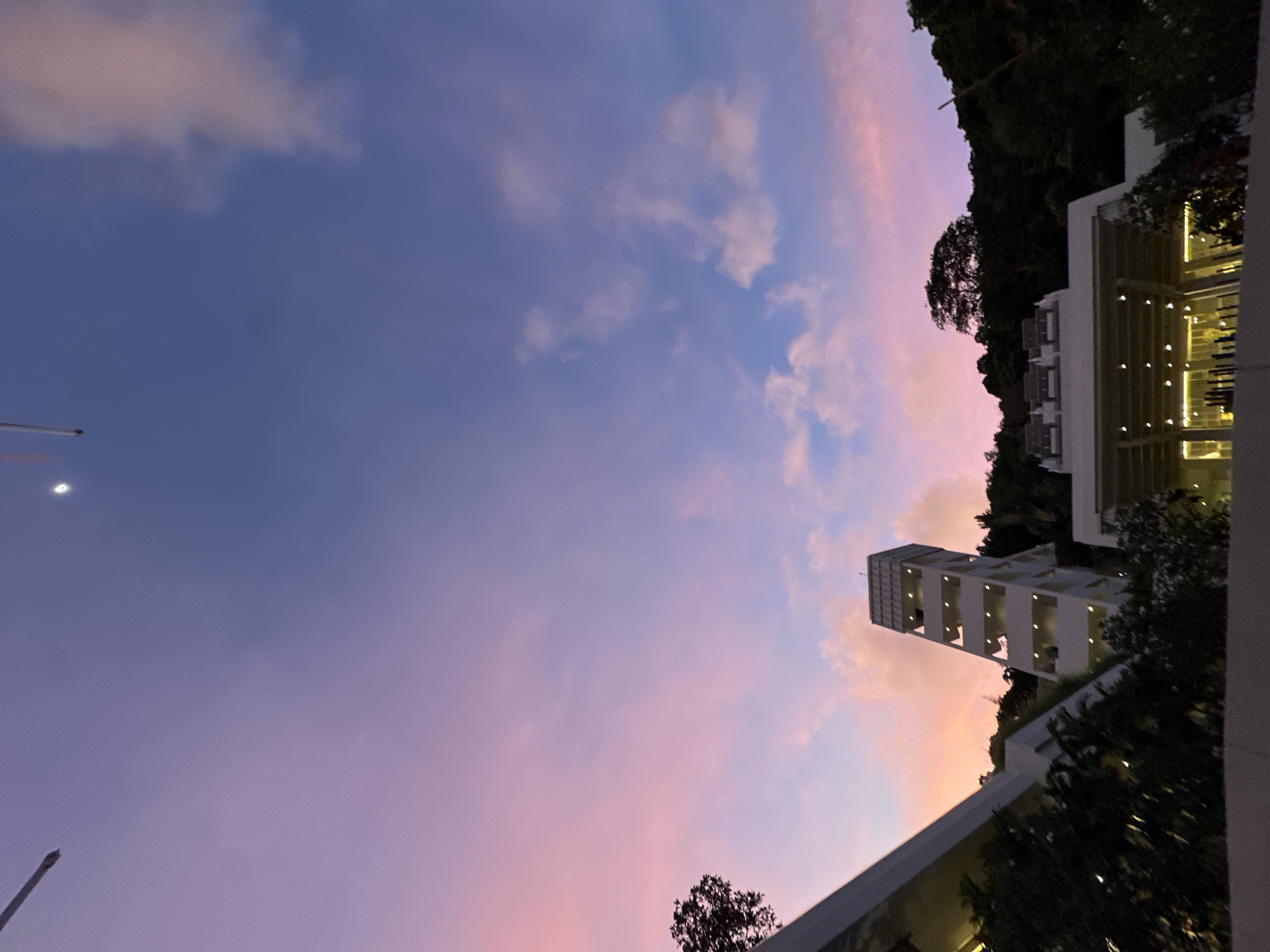 This is a wide-angle, low-angle shot taken at dusk in Rawai, Thailand. The sky is a gradient of soft pinks and purples, with some wispy clouds. In the foreground, the upper portion of a modern, white building with a slanted roof is visible on the left. To the right, a distinct, multi-story, white tower structure with open walkways and visible lights is situated on a hillside. Behind the tower, a lush, green hillside is covered in dense foliage and trees. The top of another building, appearing to be an open-air pavilion with seating, is visible on the right side of the frame. The lighting suggests it is twilight, with artificial lights beginning to illuminate the structures.