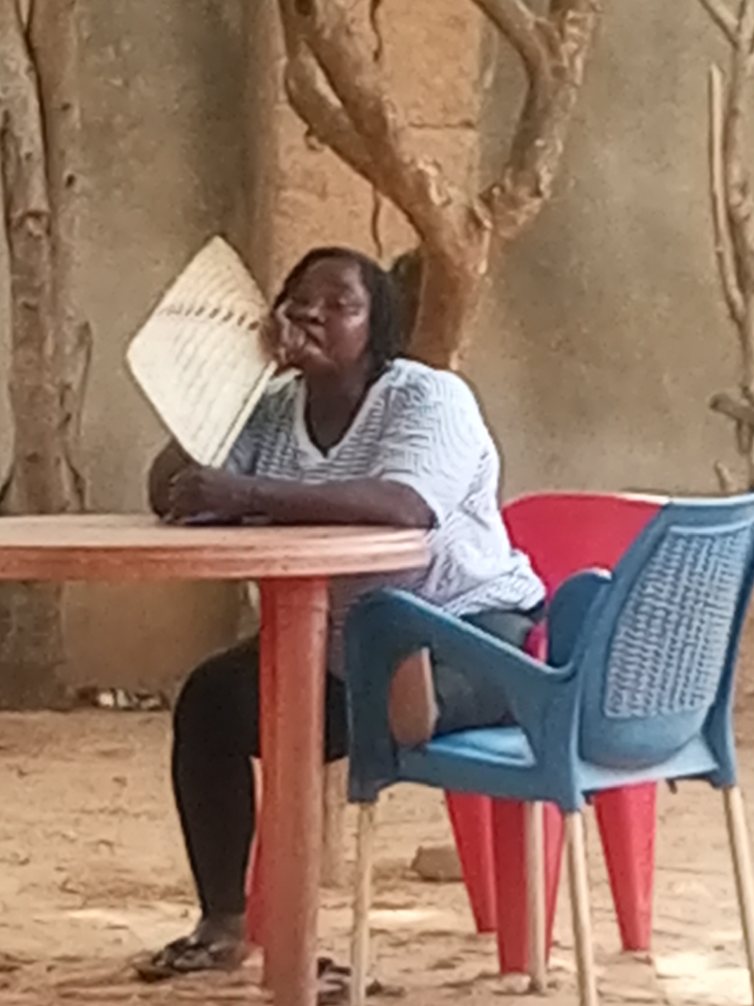 A person is sitting at a round, red table outdoors in Unknown, Unknown. The individual, wearing a light-colored, striped shirt and dark pants, is seated in a blue plastic chair with a red chair behind it. The person holds a woven fan up to their face. A tree with bare branches is visible in the background. The ground is sandy.