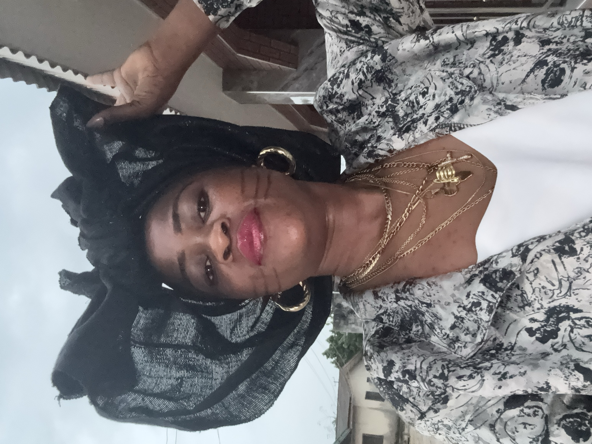 A woman is pictured outdoors in Ajara, Nigeria, wearing a black headwrap, gold hoop earrings, and a patterned jacket over a white top. She is adorned with multiple gold necklaces, one of which features a cross pendant. The background shows residential buildings and overcast skies. The woman is holding her headwrap with her right hand.