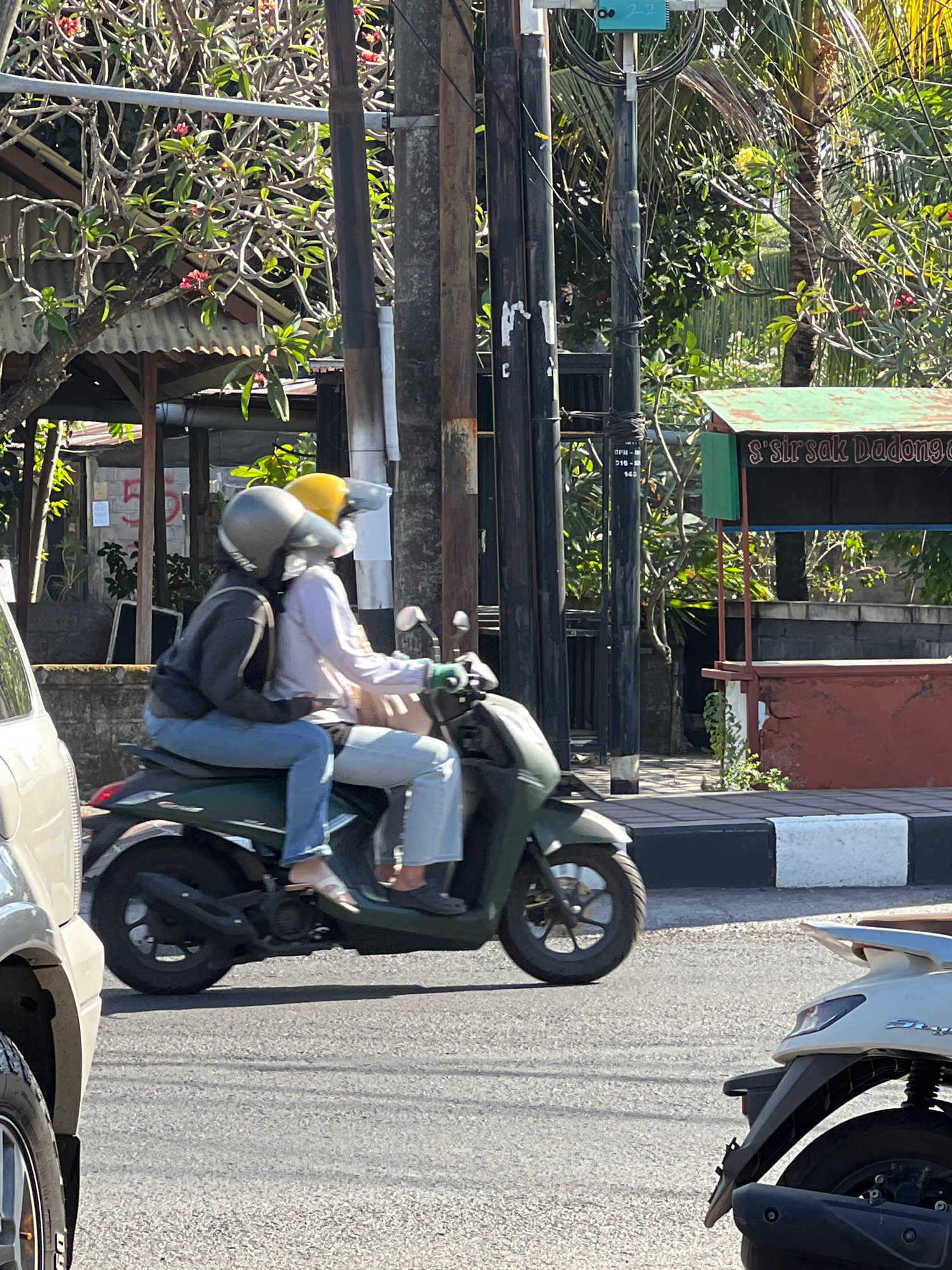 In Denpasar, Indonesia, two individuals are riding a dark green scooter. The person in front, wearing a dark grey hoodie and blue jeans, is driving. The passenger behind, in a light pink long-sleeved shirt and light blue jeans, has their arms around the driver. Both are wearing helmets, the driver's is dark grey, and the passenger's is yellow. The scooter is in motion on a paved road, with a white vehicle to the left and another light-colored scooter to the right. In the background, there are trees with red and yellow flowers, utility poles with many wires, a weathered shed with a corrugated metal roof, and a small food stall with a green awning and a sign that reads "S'sir sak Dadong". The scene is illuminated by bright sunlight.