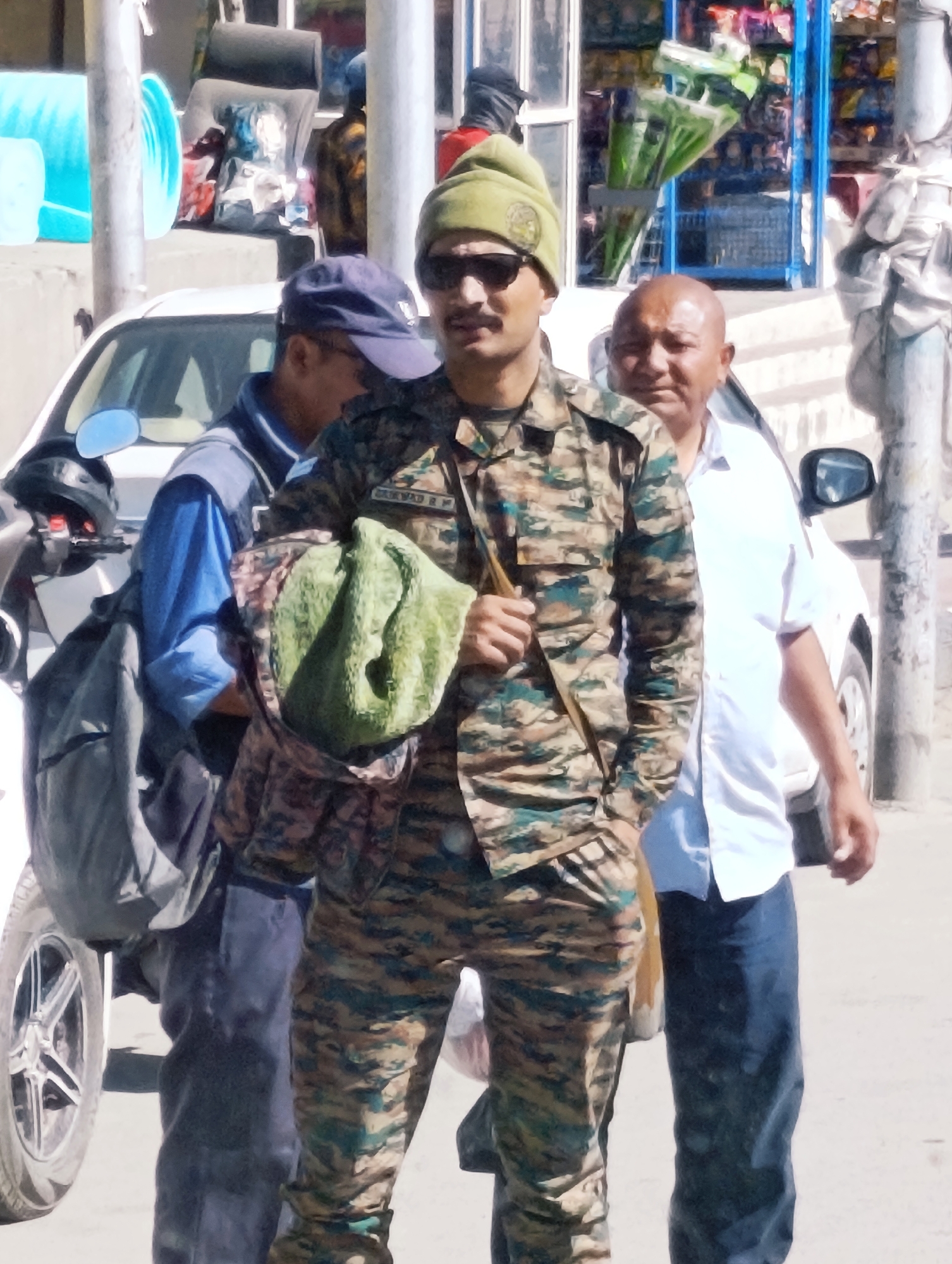 In Leh, India, three men are depicted on a paved surface, possibly a street. A man in the foreground, wearing a camouflage uniform and a green beanie, stands facing the camera. He has a mustache and sunglasses. He holds a green cloth in his left arm, which is partially tucked into his uniform. To his left, a man in a blue shirt and dark pants, wearing a dark blue cap, is partially visible. He appears to be looking down. To the right of the man in camouflage, another man wearing a white shirt and dark pants is visible, looking towards the camera. A white car is parked behind the men, with a portion of its wheel visible in the foreground. A motorcycle with a helmet is parked to the left of the man in the blue shirt. In the background, a shop is visible with shelves stocked with various items. A metal pole stands to the right of the frame.