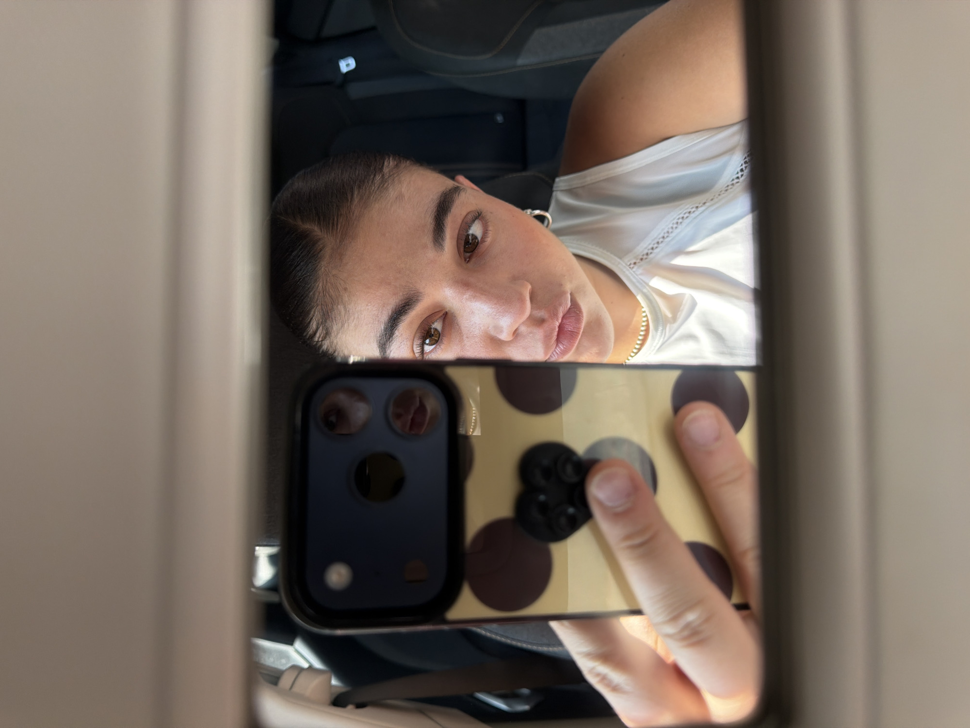 A person is reflected in a mirror, holding a cell phone with a polka-dot case. The person has light skin, dark hair pulled back, and is wearing a white shirt with lace trim. A necklace is visible around their neck. The mirror is surrounded by off-white trim. The scene appears to be inside a car, possibly in the city of Puebla, Mexico, though there are no specific visual clues to confirm the location. The focus is on the reflection of the person and the phone.