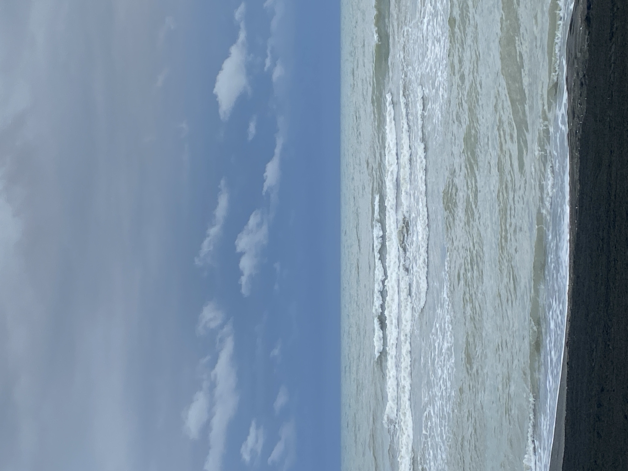 The image shows a view of the Black Sea coast in Ureki, Georgia. The sky is overcast with dark blue and grey clouds, with some lighter white clouds scattered across the upper portion. The sea is choppy with white-capped waves rolling towards a dark-colored shore. The water appears murky and light green-grey. The beach in the foreground is dark and textured, possibly sand or pebbles. No people or structures are visible in the scene.