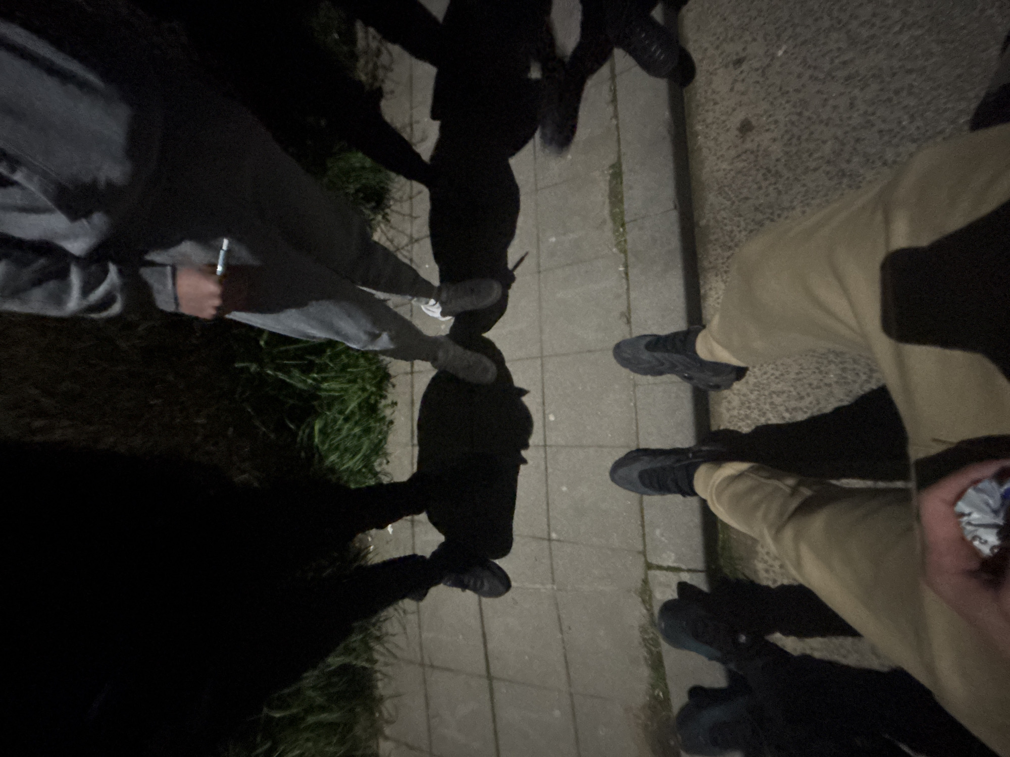 A group of people are standing together on a paved surface, likely a sidewalk, in what appears to be an urban environment. The photograph is taken from a high angle, looking down at the group. There are at least four people visible, based on the number of pairs of feet and the shadows cast. One person is wearing gray sweatpants and gray sneakers, holding a metallic object in their hand. Another person is wearing tan pants and dark-colored sneakers. Others are wearing dark clothes with black shoes. The location of the image is unknown. There is grass visible at the edge of the pavement, suggesting a park or residential area. The overall lighting is poor, likely taken at night or in a dimly lit area.