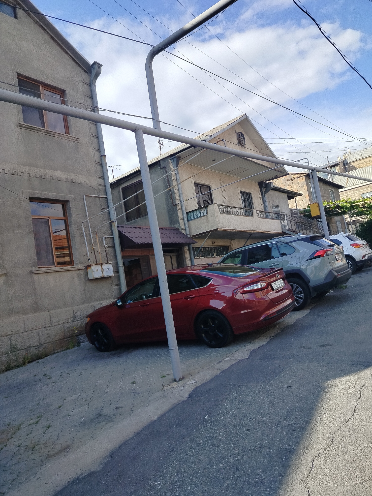 The image depicts a street scene in Yerevan, Armenia, characterized by parked cars along a narrow, sloping road flanked by buildings with varied architectural styles. A red Ford Fusion is parked adjacent to a weathered, stone building with two visible windows. Beside the Fusion is a grey Toyota RAV4, and further up the street is a white compact SUV. Overhead, a network of utility poles and wires crisscrosses the sky, and the buildings are close together, typical of urban settings. The sky is partly cloudy. The road surface is asphalt, and the sidewalk is paved with stones.