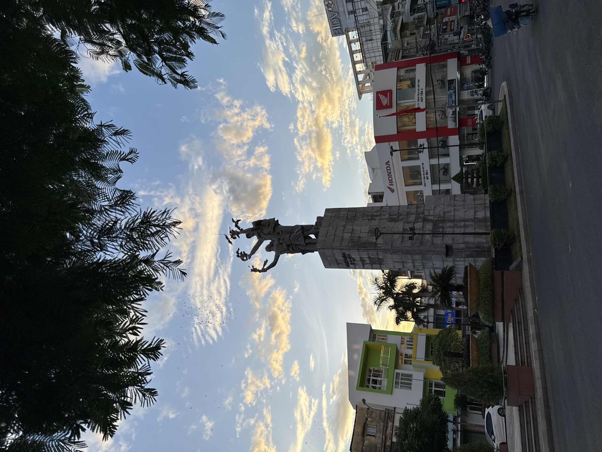 This image depicts a street view in Nha Trang, Vietnam, featuring a prominent monument in the foreground, silhouetted against a sky filled with scattered clouds. The monument, appearing to be made of stone, has a statue of figures at its apex, holding aloft what seem to be birds or branches.
To the left of the monument, a modern building with a striking green and white facade stands out, with multiple windows and balconies. Adjacent to it, older, more weathered buildings are visible. In the background, a row of shops and businesses line the street, with one clearly displaying the "HONDA" logo on its red and white storefront. Further down the street, other commercial buildings and a BIDV bank are identifiable.
The street itself is paved, with a sidewalk or plaza area in front of the monument, featuring planters with small trees and bushes. Several cars are parked or driving on the street. A person on a motorcycle, laden with cargo and wearing a conical hat, is seen riding away from the camera on the right side of the image. The overall lighting suggests it is either early morning or late afternoon, with the sun casting a warm glow on the clouds. Lush trees, including palm trees, are also present, adding to the tropical atmosphere. A flock of small birds is scattered across the sky.
