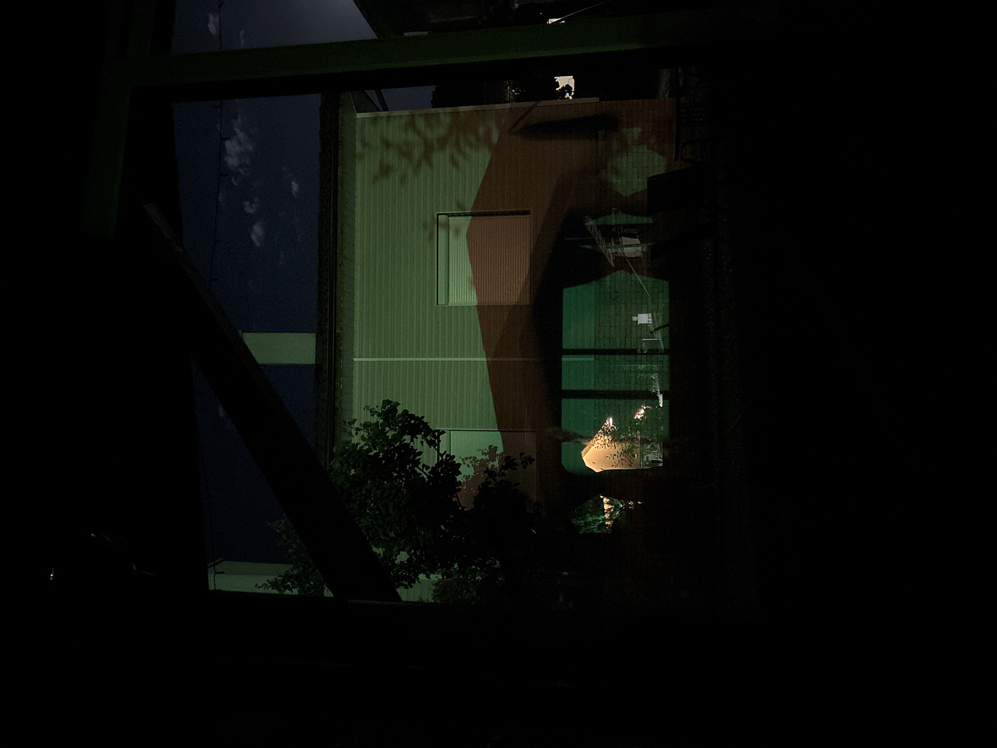 The image shows a view from inside a dark space, framed by a dark green structure, likely part of a building or industrial framework. Through this opening, a building with vertical siding is visible, illuminated by artificial light. The building's facade appears to have a window and is partially covered by shadows, possibly cast by tree branches. To the left, a dark blue area suggests a night sky. The lower part of the view is obstructed by dark foliage. The city of Polena, Bulgaria is not discernible in the image.