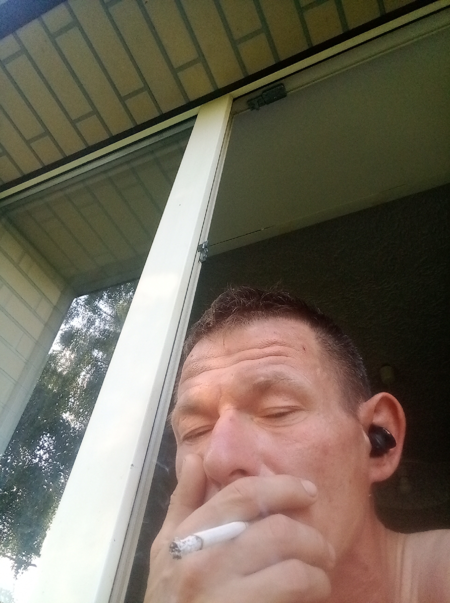 A close-up, high-angle, outdoor selfie shows a white man with closed eyes holding a lit cigarette to his mouth with his right hand, the ash end visible. He has short brown hair, wrinkles on his forehead, and a black wireless earbud in his right ear. He is wearing a light-colored sleeveless shirt. The background includes a white window frame, a window with a reflection of trees and sky, and a tiled overhang. The image appears to have been taken from a window or doorway. No specific location in Tapa, Estonia is identifiable.