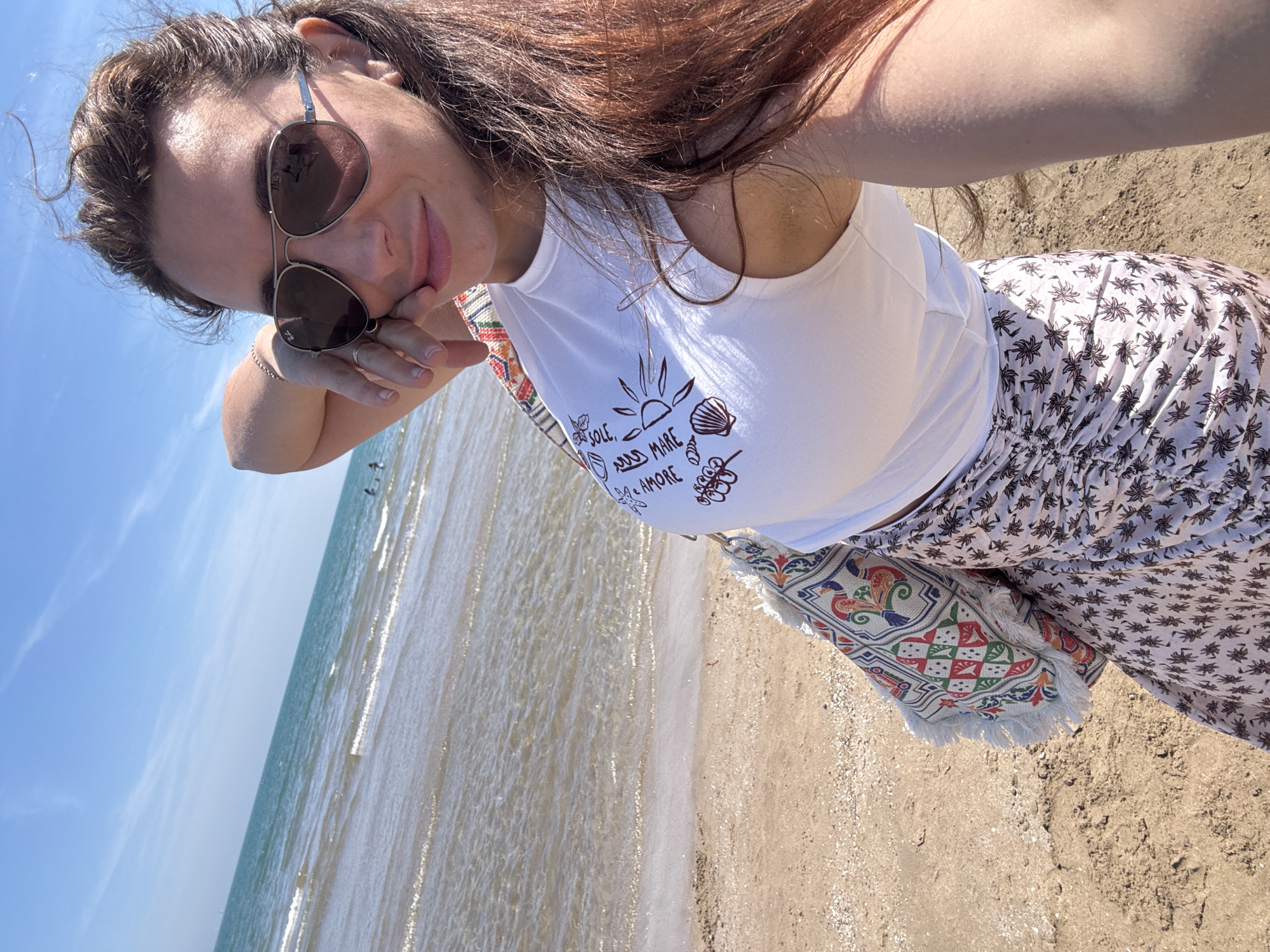 A woman is standing on a sandy beach adjacent to a calm sea under a clear blue sky. She wears sunglasses, a white tank top with a brown print on it, and pants with a palm tree print. The woman's right hand is touching her face. A bag with a patterned design and fringe is slung over her shoulder. The location is likely a beach in Valencia, Spain, given the typical beach and sea characteristics of that region.