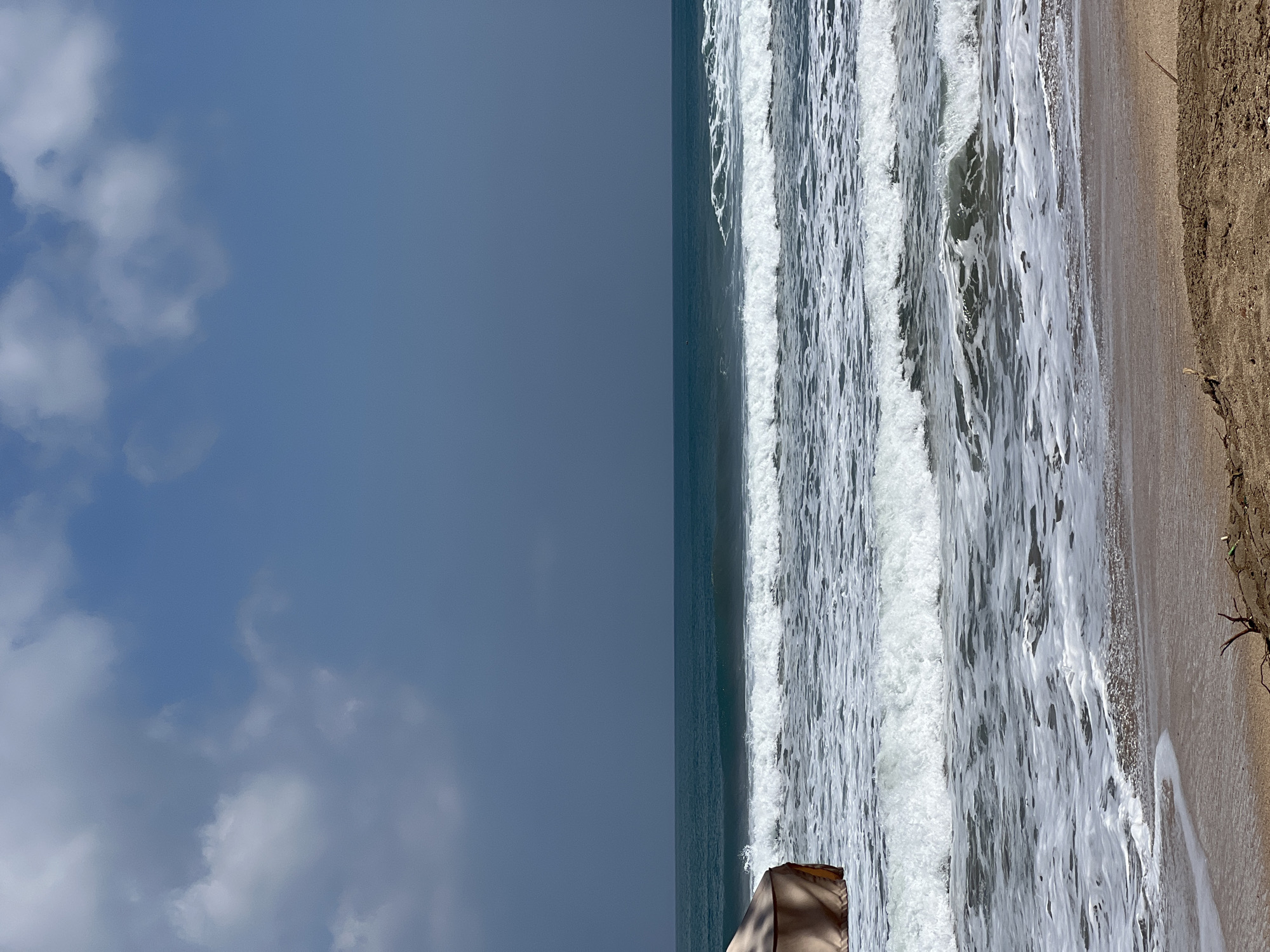 A beach scene in Kuta, Indonesia, depicts a sandy shore in the foreground leading to the ocean. Several waves are breaking, creating white foam and churned water as they approach the beach. The ocean is a deep blue-green color, with a clear horizon line separating it from a hazy blue sky filled with scattered white clouds. On the left side of the frame, a portion of a light brown beach umbrella or canopy is visible, casting a shadow on the sand. The overall environment suggests a tropical beach setting, possibly a cloudy day.