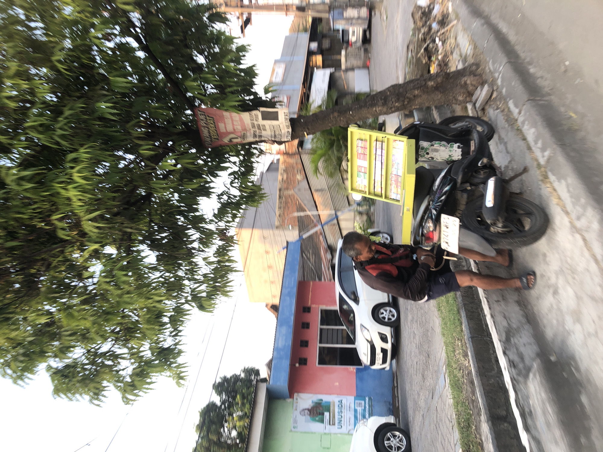 In Tenggulunan, Indonesia, a man wearing a dark long-sleeved jacket with red accents and dark shorts stands beside a black motorcycle. The motorcycle has a yellow display rack attached to its rear, filled with various cigarette packs. The man appears to be interacting with the motorcycle or the display. The scene is on a street with a sidewalk. In the background, there are buildings, including one with a blue roof and a pink facade, and another with a green facade displaying a poster. White cars are parked on the street. A tree with dense green foliage stands to the right of the motorcycle, with a sign attached to its trunk. The lighting suggests it is daytime, possibly late afternoon due to the angle of the shadows.