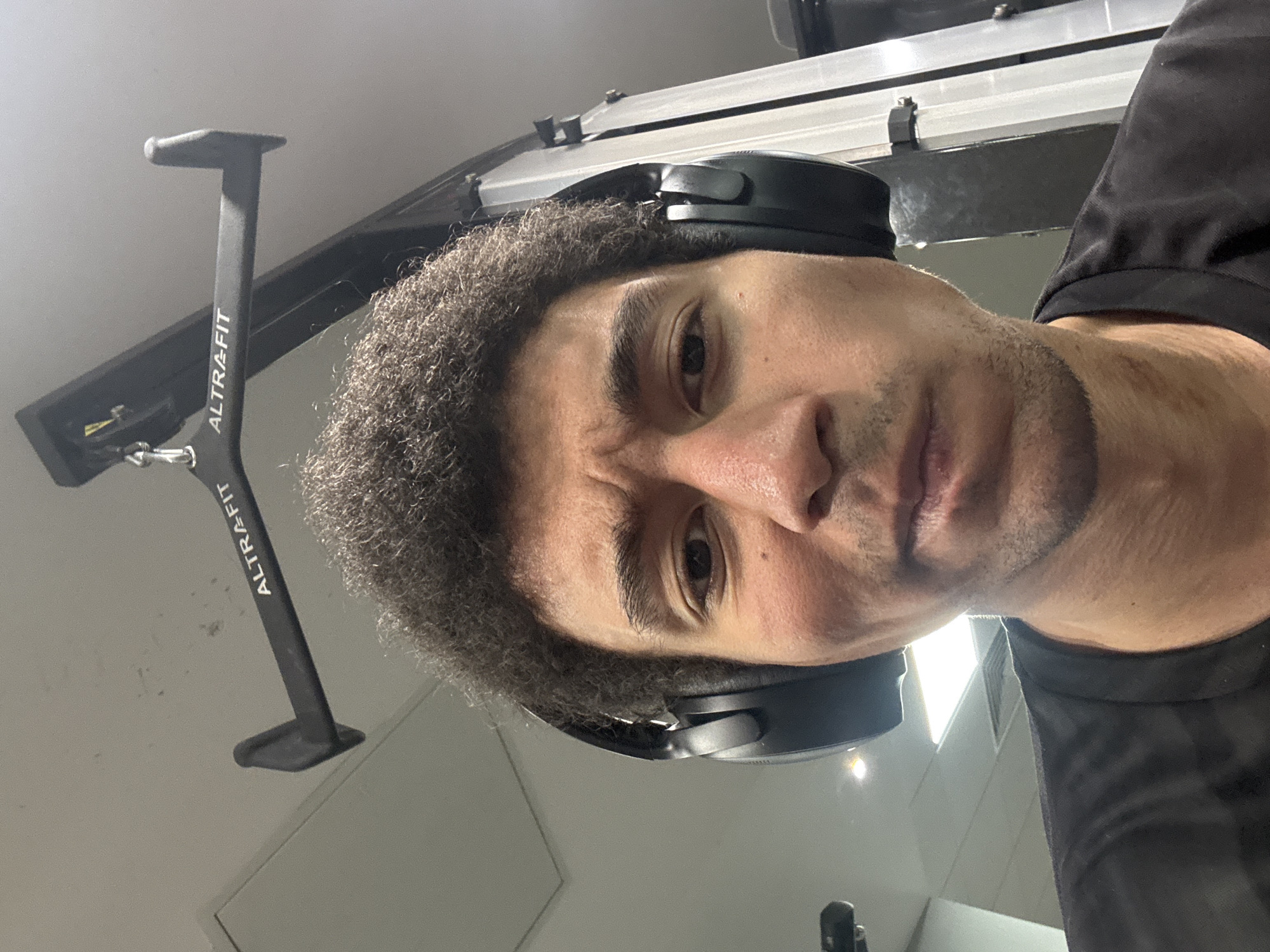 The image is a close-up, rotated selfie of a man, taken indoors, likely in a gym. The man, with dark, curly hair and stubble, wears black over-ear headphones and a dark t-shirt. He looks directly at the camera with a serious and slightly intense expression, his eyebrows furrowed, creating visible wrinkles on his forehead.
In the background, to the left of the man, a black piece of gym equipment is visible. The brand name "ALTRAFIT" is clearly printed vertically in white text on its stand. Other background elements include metallic structures, possibly part of gym machinery or ceiling fixtures, and artificial lighting reflecting off surfaces, confirming an indoor setting. The low angle of the shot and the man's direct gaze suggest he is taking a self-portrait. The scene is consistent with a workout environment in a gym, specifically located in London, United Kingdom, as per the provided context.