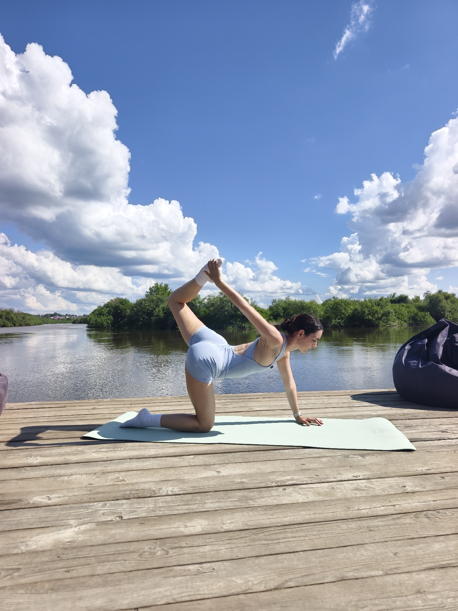 A woman is performing a yoga pose on a light green mat placed on a wooden pier. She is on her knees, with her right leg bent and her right hand reaching back to hold her right foot. Her left arm is extended forward, with her left hand flat on the mat. She is wearing a light blue athletic set and white socks.
The pier is situated next to a body of water, likely a river, with a lush green tree line on the opposite bank. The sky is blue with scattered white cumulus clouds. In the background, to the right of the woman, a dark blue beanbag chair is visible. A small portion of what appears to be a purple object is visible on the far left edge of the frame.
The scene is outdoors, suggesting a recreational area or a natural setting. The overall impression is one of tranquility and physical activity in a serene environment. There is no indication of the city of Онохино, Russia, in the image itself.