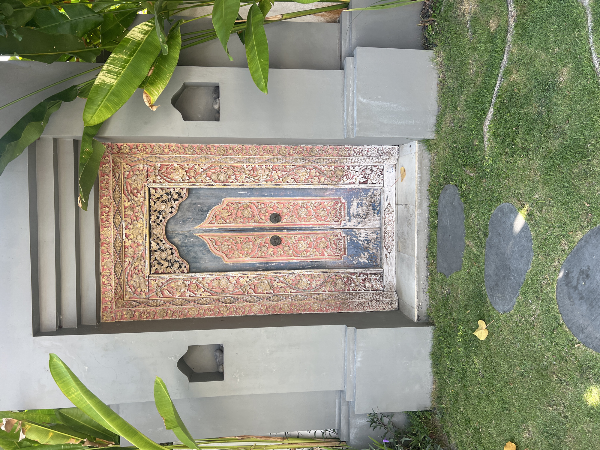 The image depicts an ornate, double-leaf wooden door, intricately carved and painted in shades of red, gold, and dark blue. The door is set within a modern, grey concrete structure that features two small, arched niches on either side, each containing a few stones. Tall, lush green banana leaves frame the sides of the door and the concrete structure. In front of the door, a pathway of irregularly shaped dark grey stepping stones is laid across a neatly mown green lawn. A few fallen yellow leaves are scattered on the grass. The scene appears to be an entrance to a building in a tropical setting, likely a resort or private residence in North Kuta, Indonesia, given the style of the door and the surrounding vegetation. There are no people or animals visible in the image.