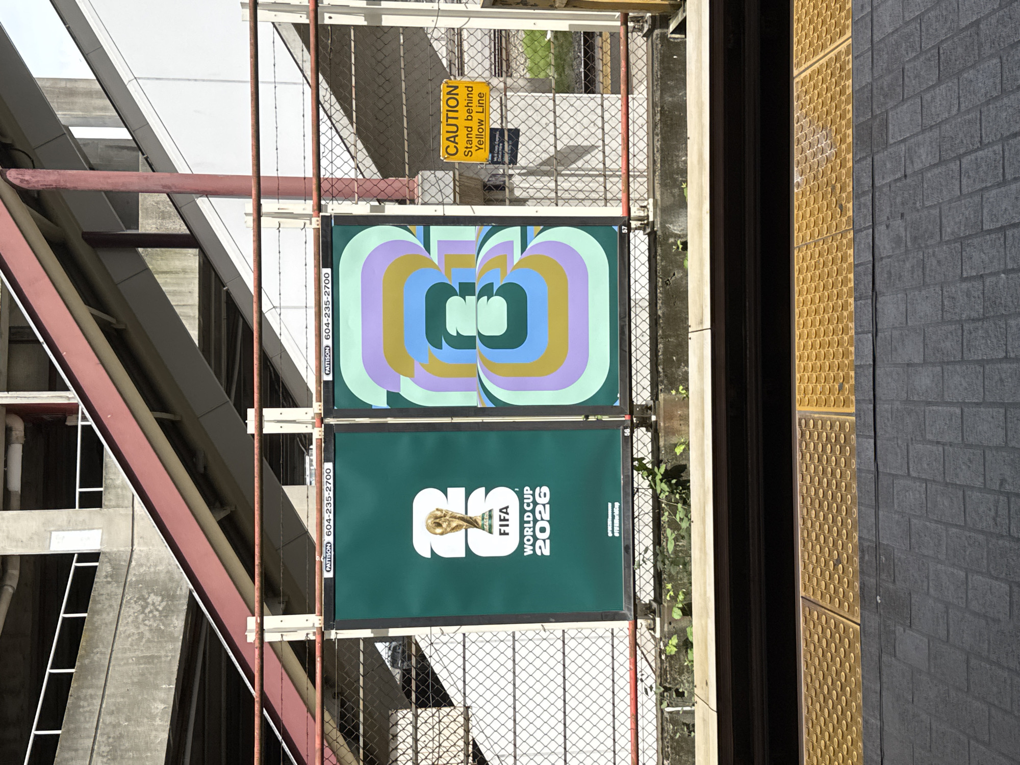 The image depicts two posters affixed to a metal fence at a transit station in Vancouver, Canada. The bottom poster advertises the FIFA World Cup 2026. Above it is a poster with an abstract design composed of concentric ovals in shades of green, purple, blue, and yellow. A yellow caution sign that reads "Caution: Stand behind Yellow Line" is fixed to the fence above the top poster. The fence runs parallel to a raised platform edge, which consists of a dark gray brick wall adjacent to a yellow tactile warning surface. The transit station's structural steel is visible to the left of the fence. The posters include the name "Pattison" and a phone number, "604-235-2700."
