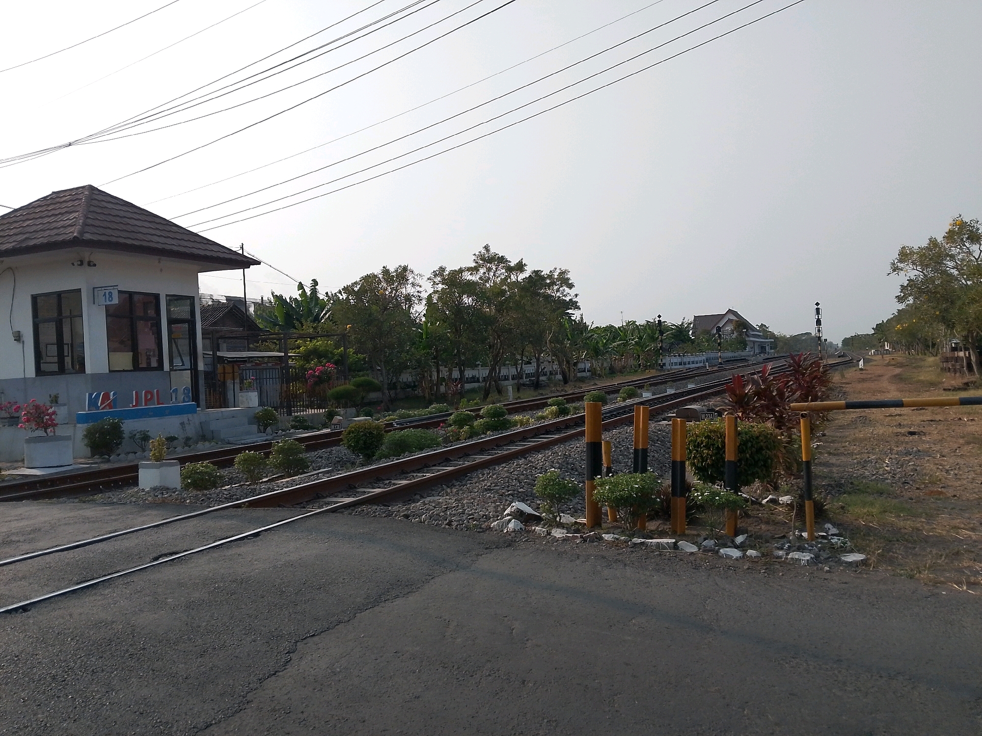 This is a medium shot of a railway crossing in Geneng, Indonesia. The image is taken at ground level, facing a railway line that runs diagonally from the bottom left to the upper right. On the left side of the frame, there is a small building with a brown tiled roof and white walls. The building has several windows and a sign that reads "KAI JPL 18". A paved road crosses the railway tracks in the foreground.
The railway tracks are lined with ballast stones and surrounded by sparse vegetation and trees. There are also several signal posts and a yellow and black barrier gate visible. The sky is clear and overcast. In the background, more trees and buildings are visible, suggesting a rural or semi-urban environment. No people are present in the image. The overall scene depicts a typical railway crossing in Indonesia during the daytime.
